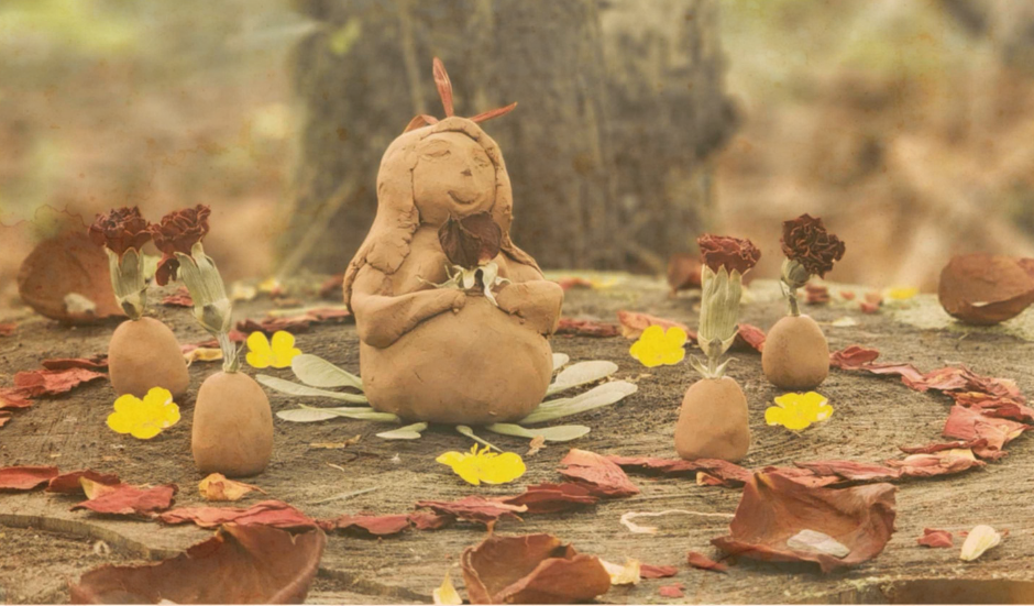 Une sculpture en argile représentant une femme avec des plumes dans les cheveux, assise sur une surface en bois avec des fleurs et des feuilles sèches décoratives autour, dans un environnement naturel.