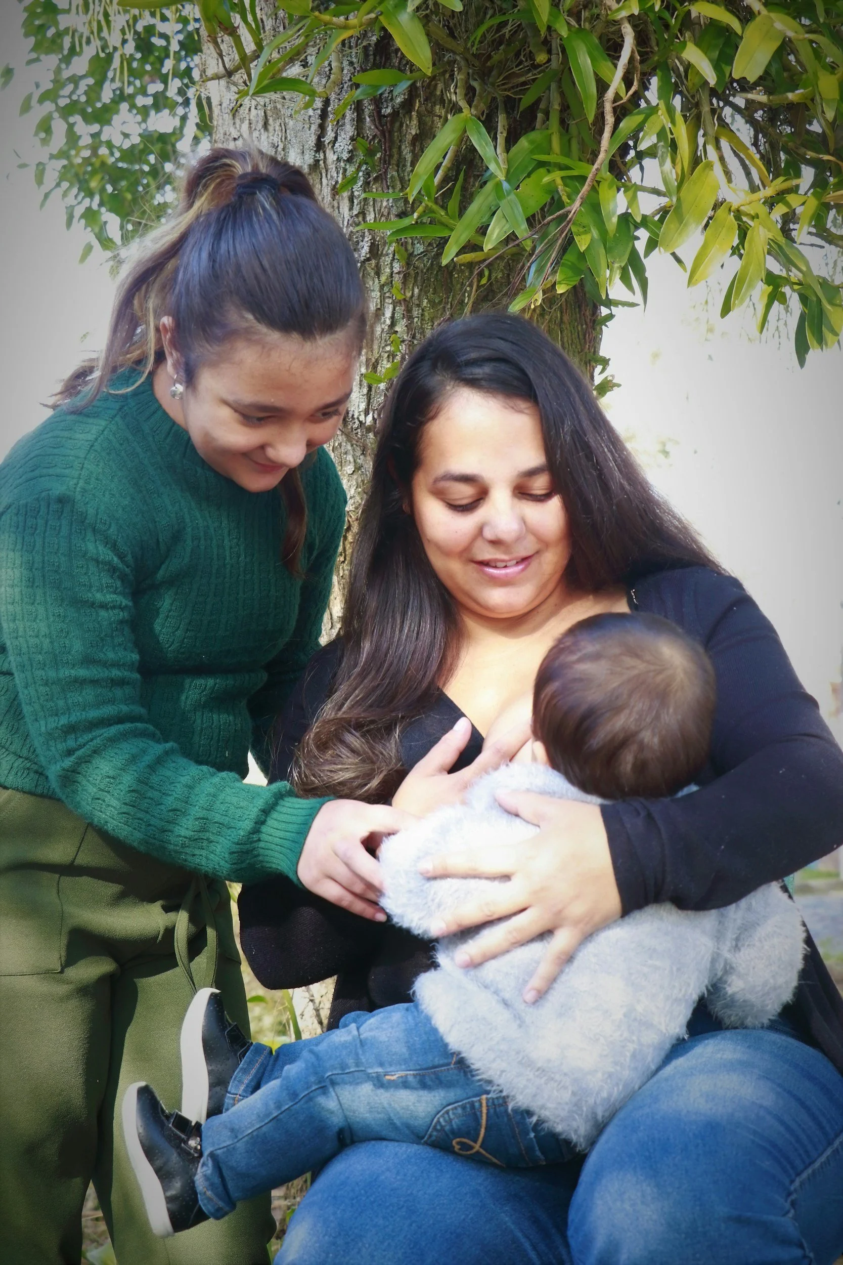 A woman breastfeeding a baby outdoors while a young girl looks on and smiles.