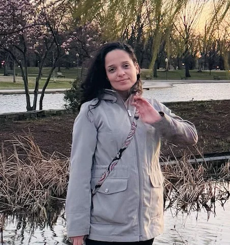 A young woman standing near a pond in a park during sunset, wearing a gray jacket and smiling at the camera.