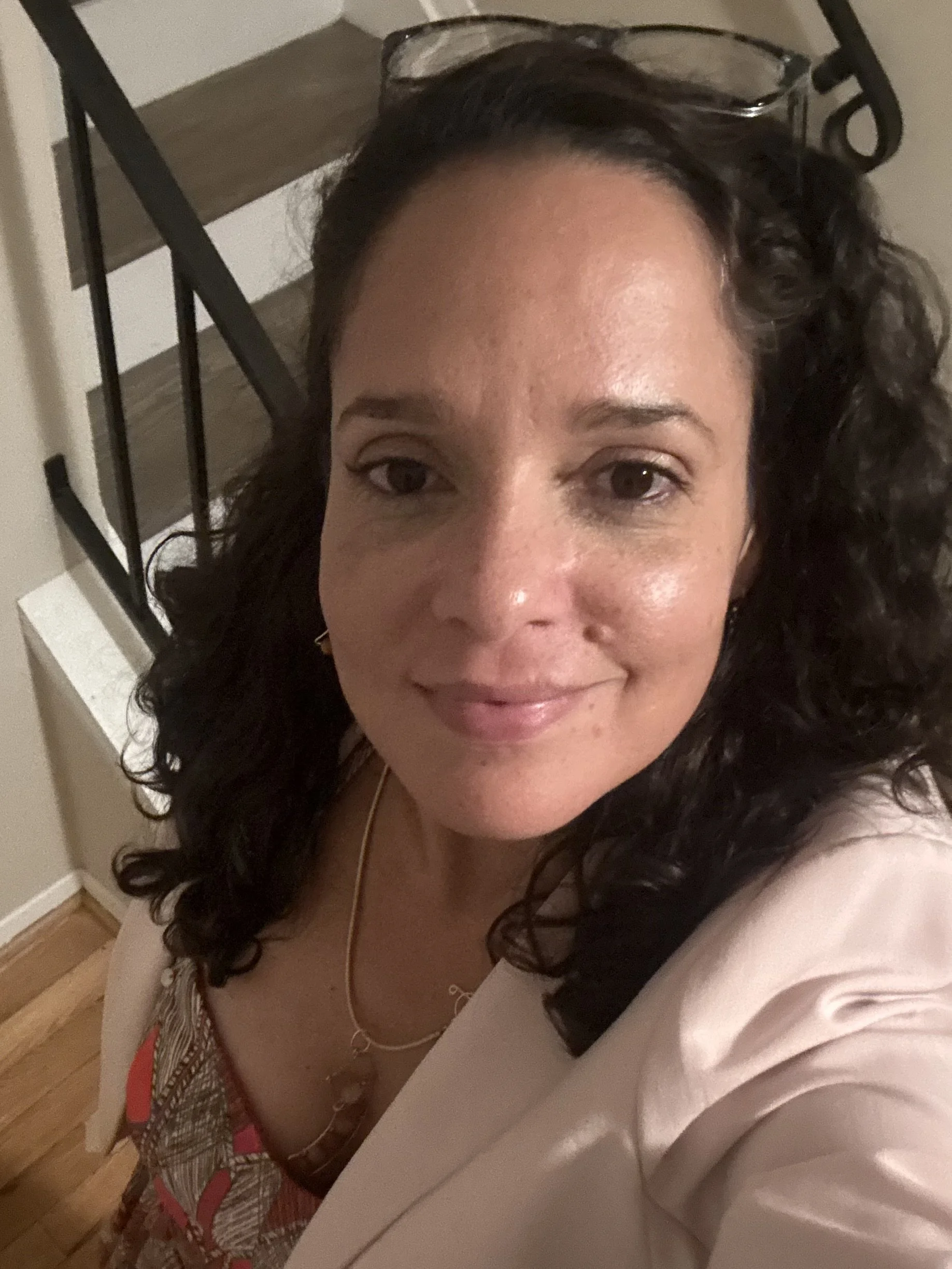 A woman with dark curly hair and glasses on top of her head, smiling, sitting at a table with a patterned purse and a red strap, in a room with wooden flooring and a white wall.
