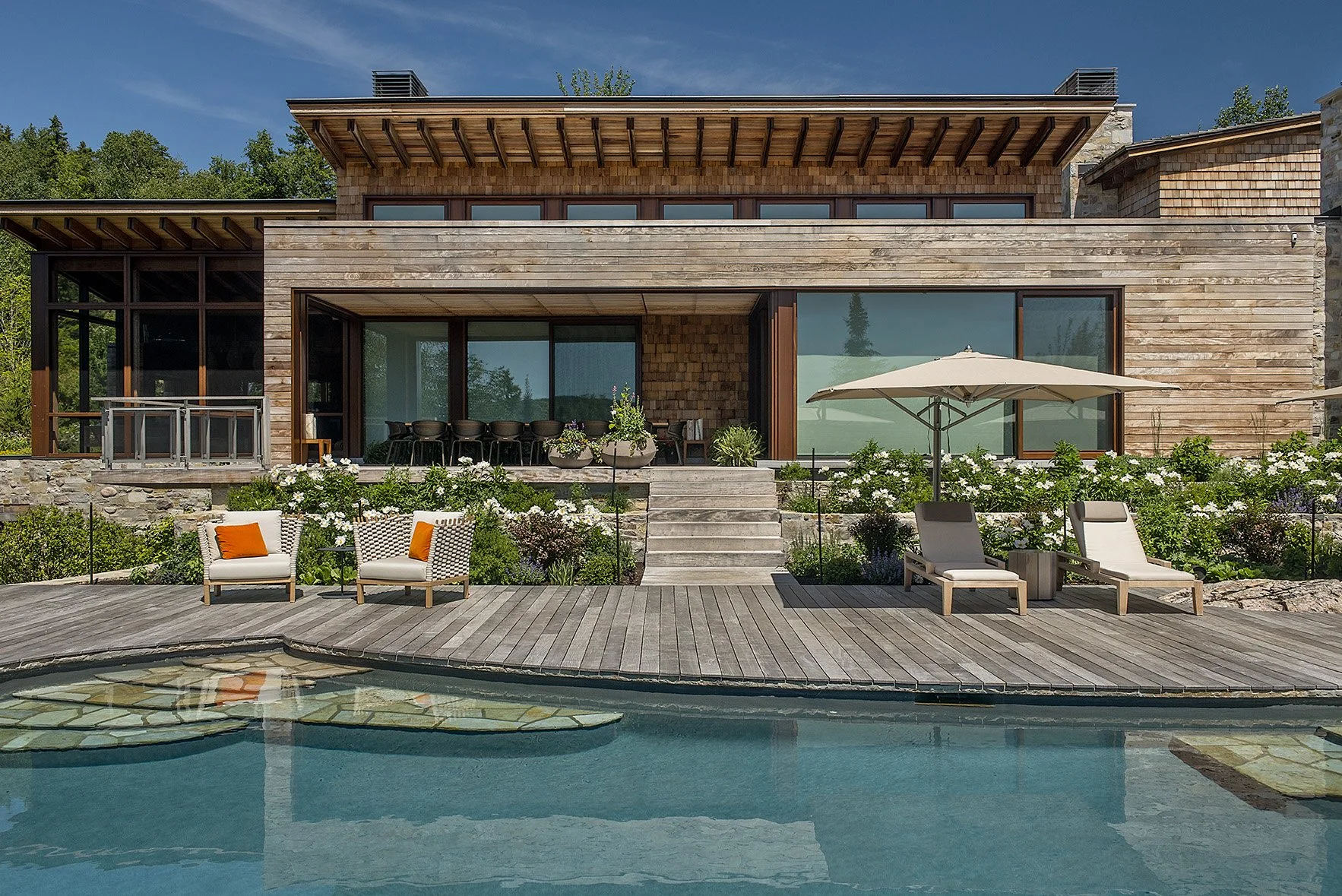 Modern house with large glass windows, wooden exterior, and outdoor patio furniture, overlooking a swimming pool with lounge chairs and umbrellas.