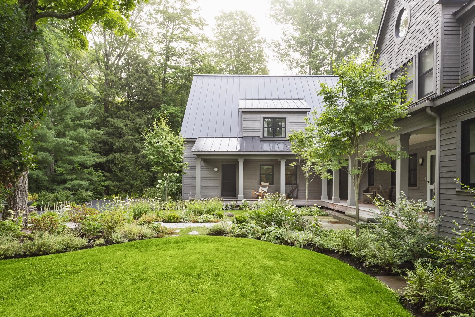 A backyard garden with green grass, trees, shrubs, and flowering plants surrounding a gray house with a covered porch and a metal roof.