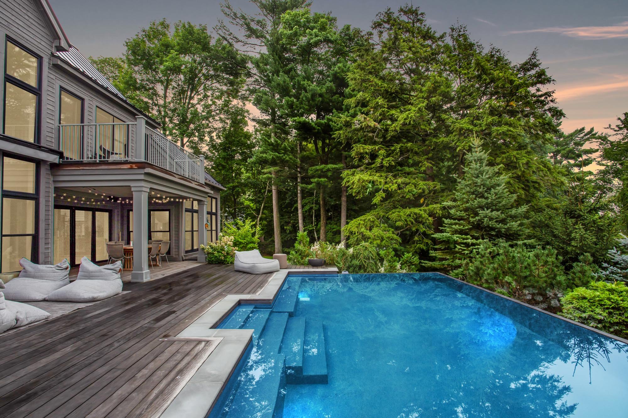 Backyard of a modern house with a wooden deck, outdoor seating, and a swimming pool surrounded by lush green trees at sunset.