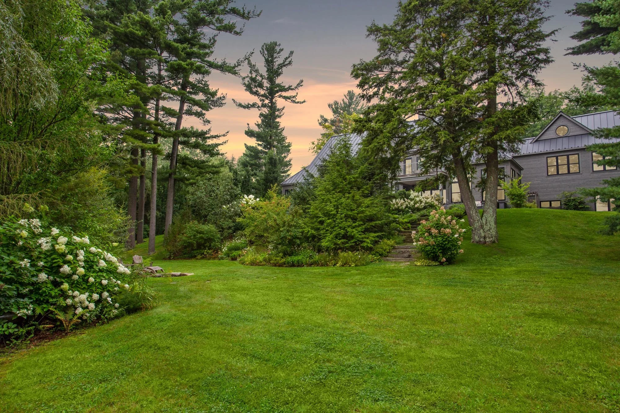 A lush green backyard with a neatly maintained lawn, surrounded by blooming bushes and tall trees, with a house with a metal roof and a staircase leading to the entrance in the background. The sky is tinged with the colors of sunset.
