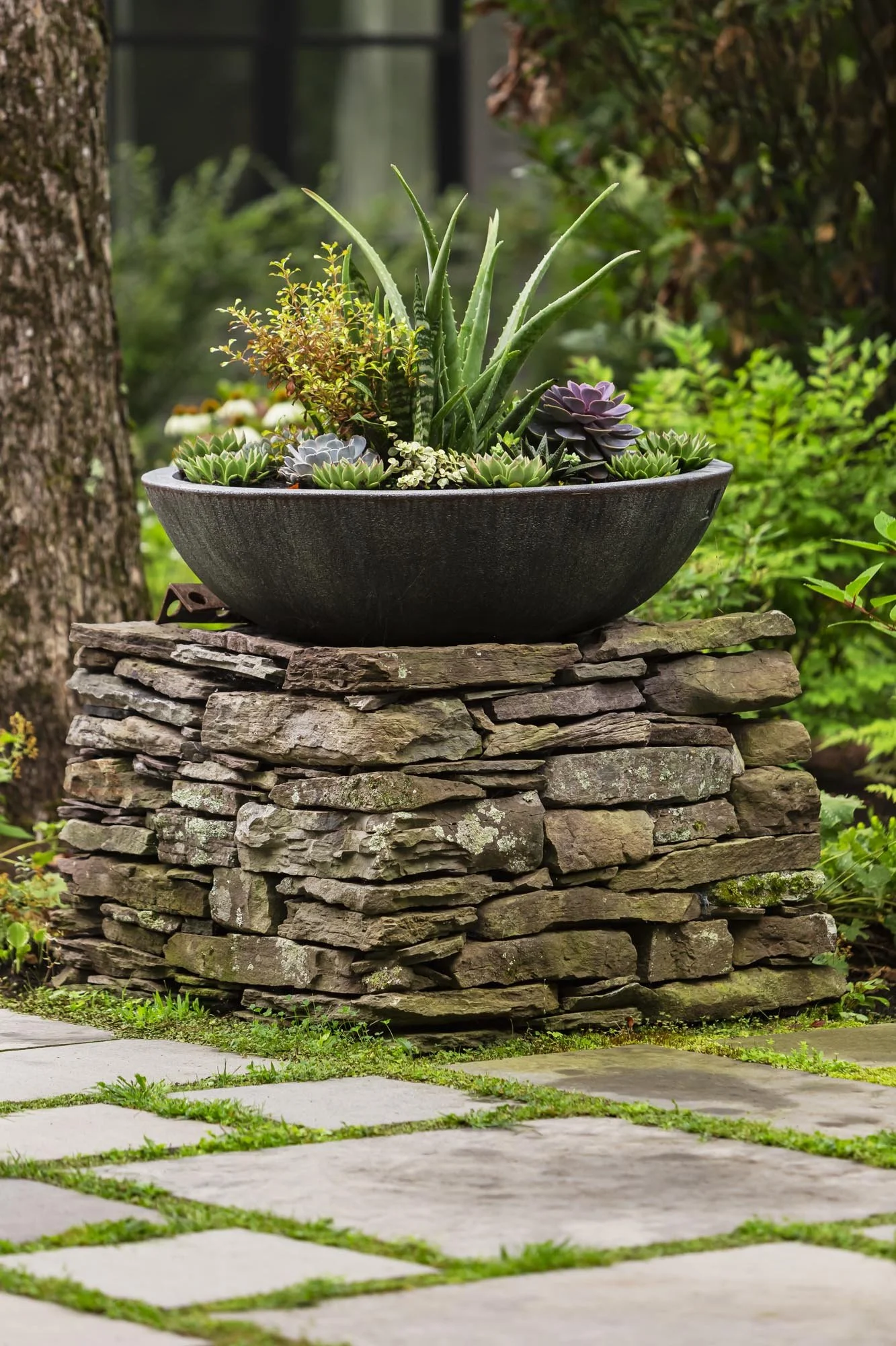 A large black planter with various succulents, including aloe, set on a stacked stone pedestal outdoors, with greenery and trees in the background.