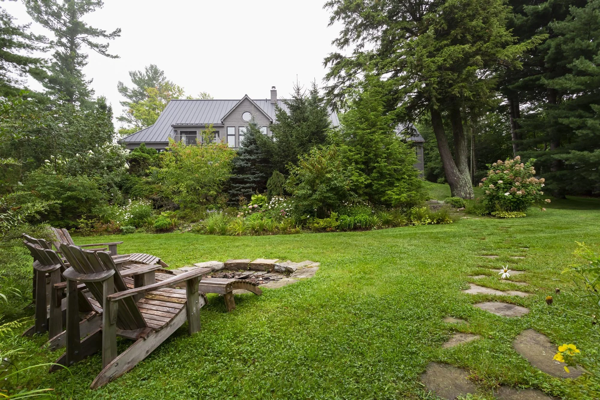 A lush green backyard with a stone pathway, wooden benches, a fire pit, and a variety of flowering shrubs and trees surrounding a gray house with a metal roof.
