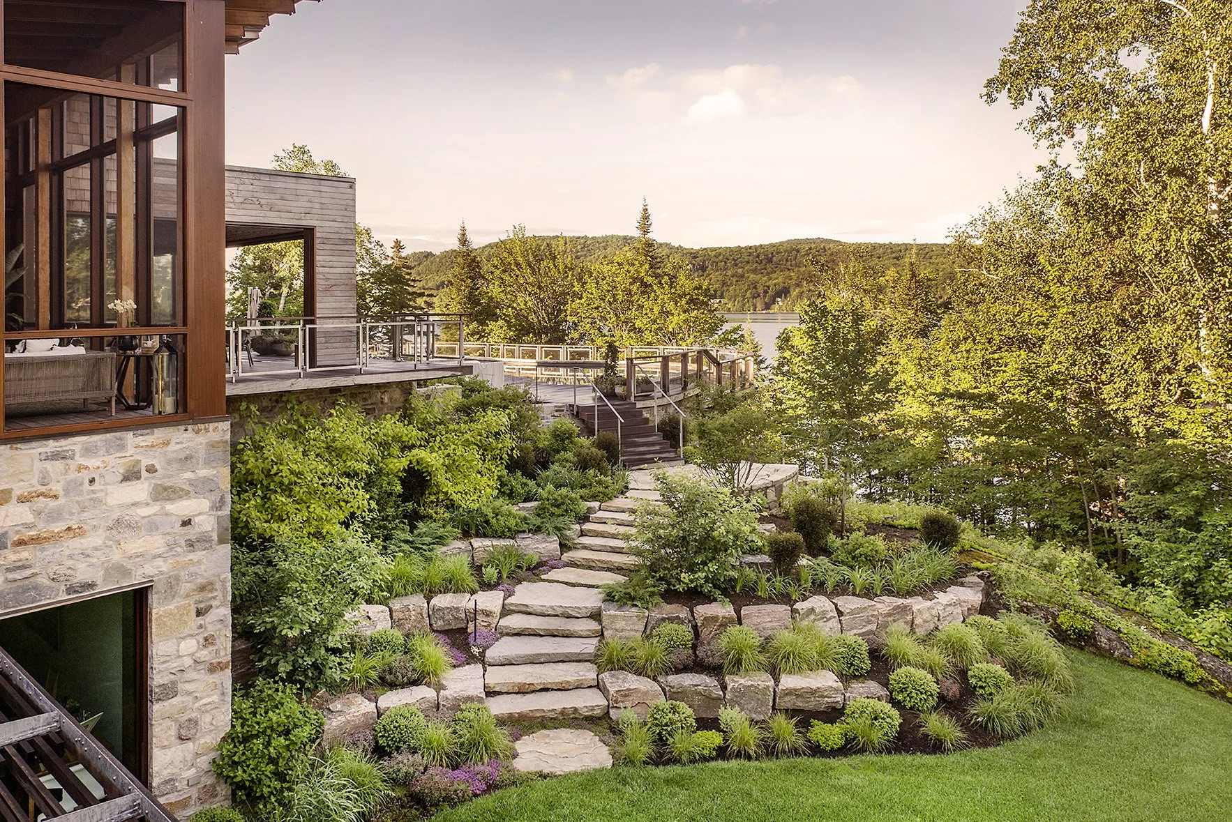 A landscaped backyard with stone steps leading down to green grass, surrounded by bushes and trees, next to a modern house with large windows and wooden accents. In the background, there is a view of a lake and distant hills under a partly cloudy sky.