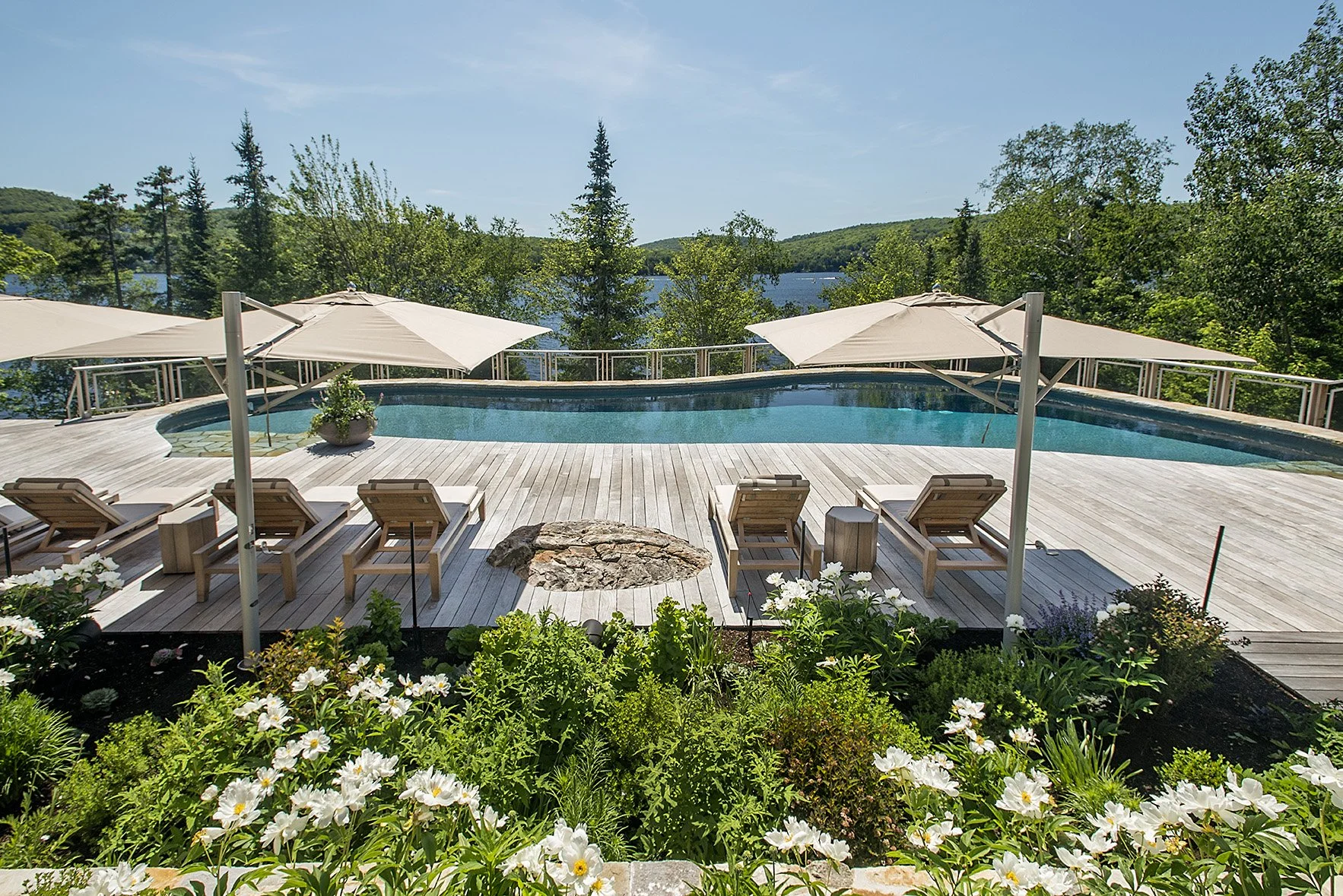 A backyard with a swimming pool, surrounded by a wooden deck and four lounge chairs with umbrellas. There are trees and a body of water in the background under a blue sky.