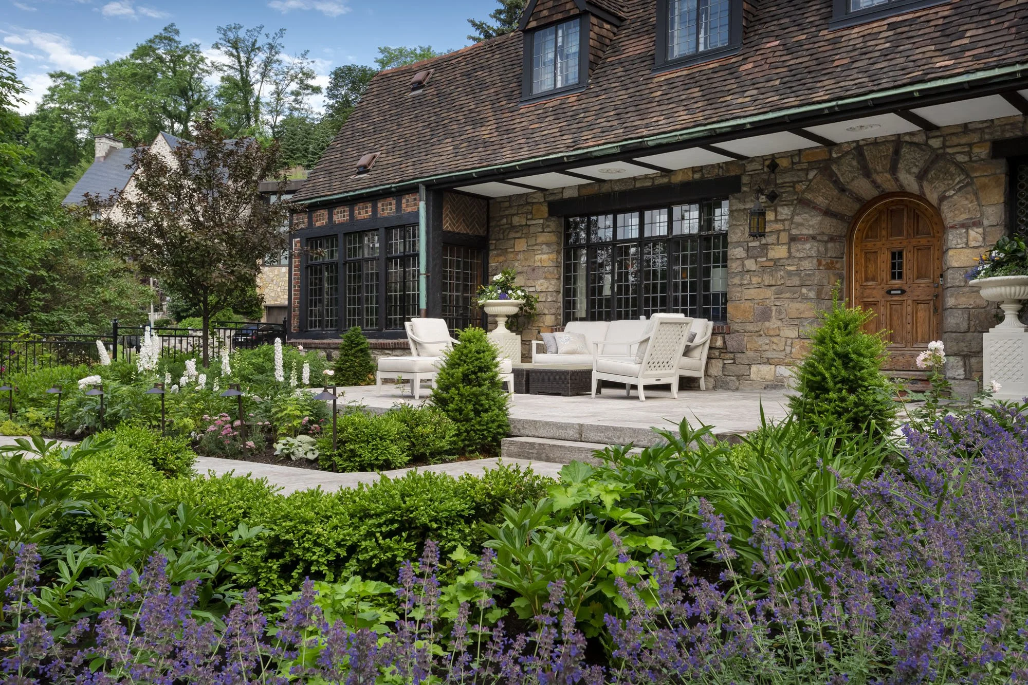 A stone house with a sloped roof and large bay windows, featuring a spacious patio with white outdoor furniture, surrounded by a lush garden with various green plants and purple flowers.