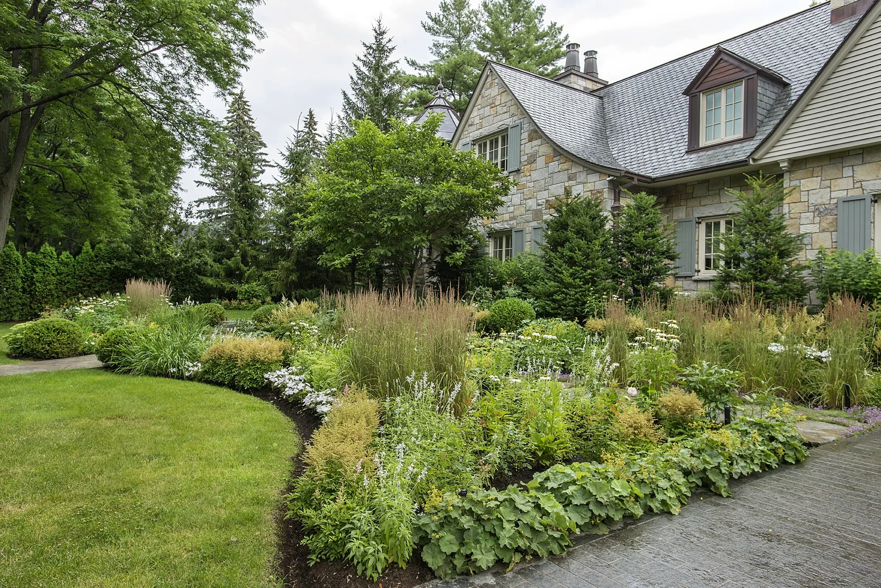 A well-maintained garden in front of a house with stone walls and gray roof, featuring a curved lawn, various flowering plants, shrubs, and tall trees.