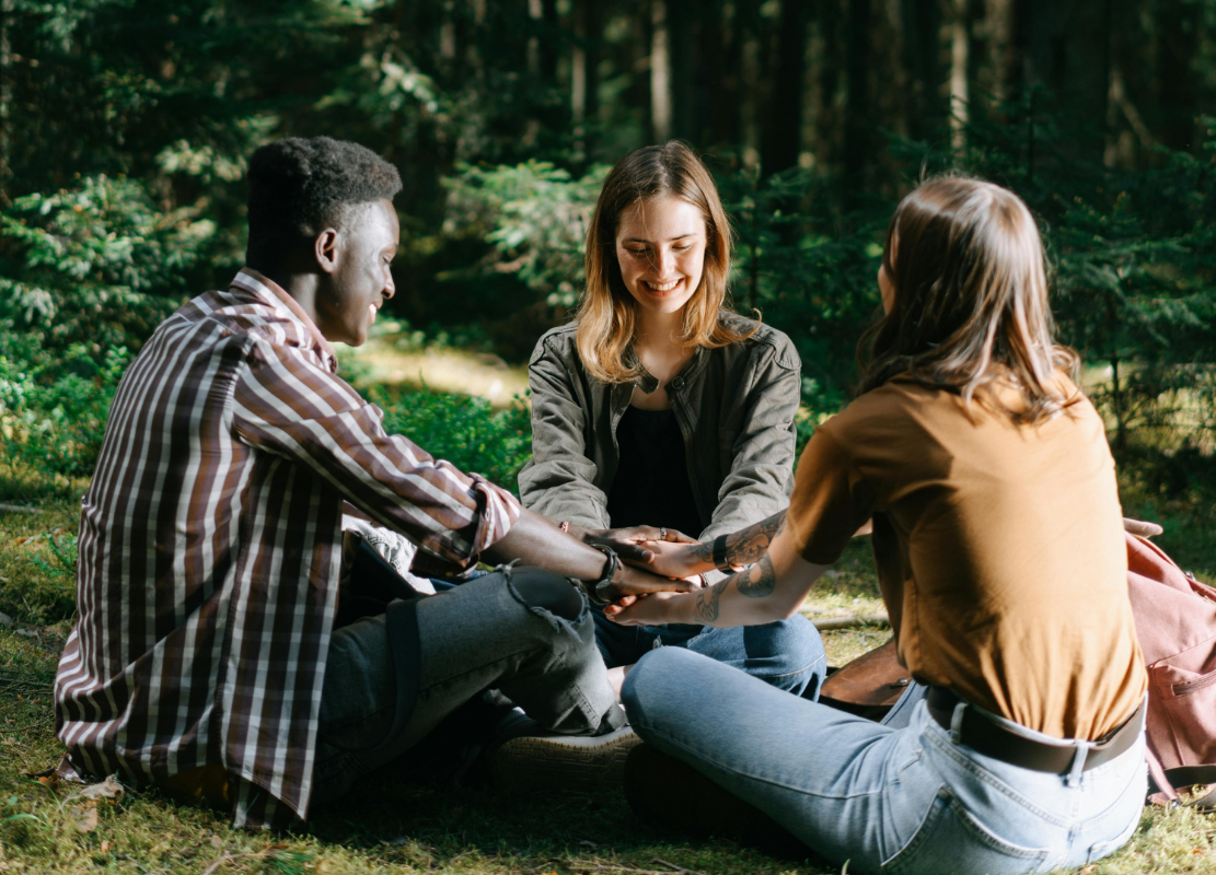 Three people sitting on grass in a forest, holding hands together, smiling and engaging in a group activity.