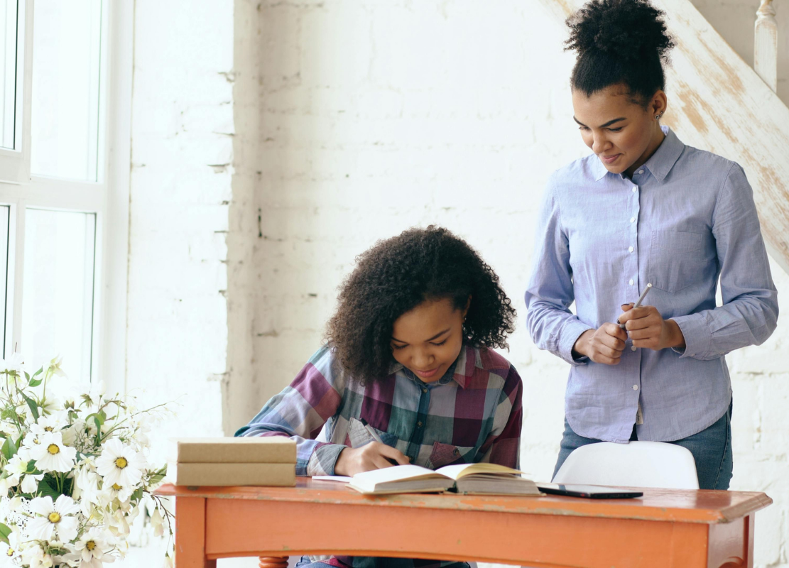 Two women working together at a desk in a bright room with white brick walls and natural light. One woman with curly hair is writing in a notebook, while the other standing woman with her hair in a bun looks on attentively.