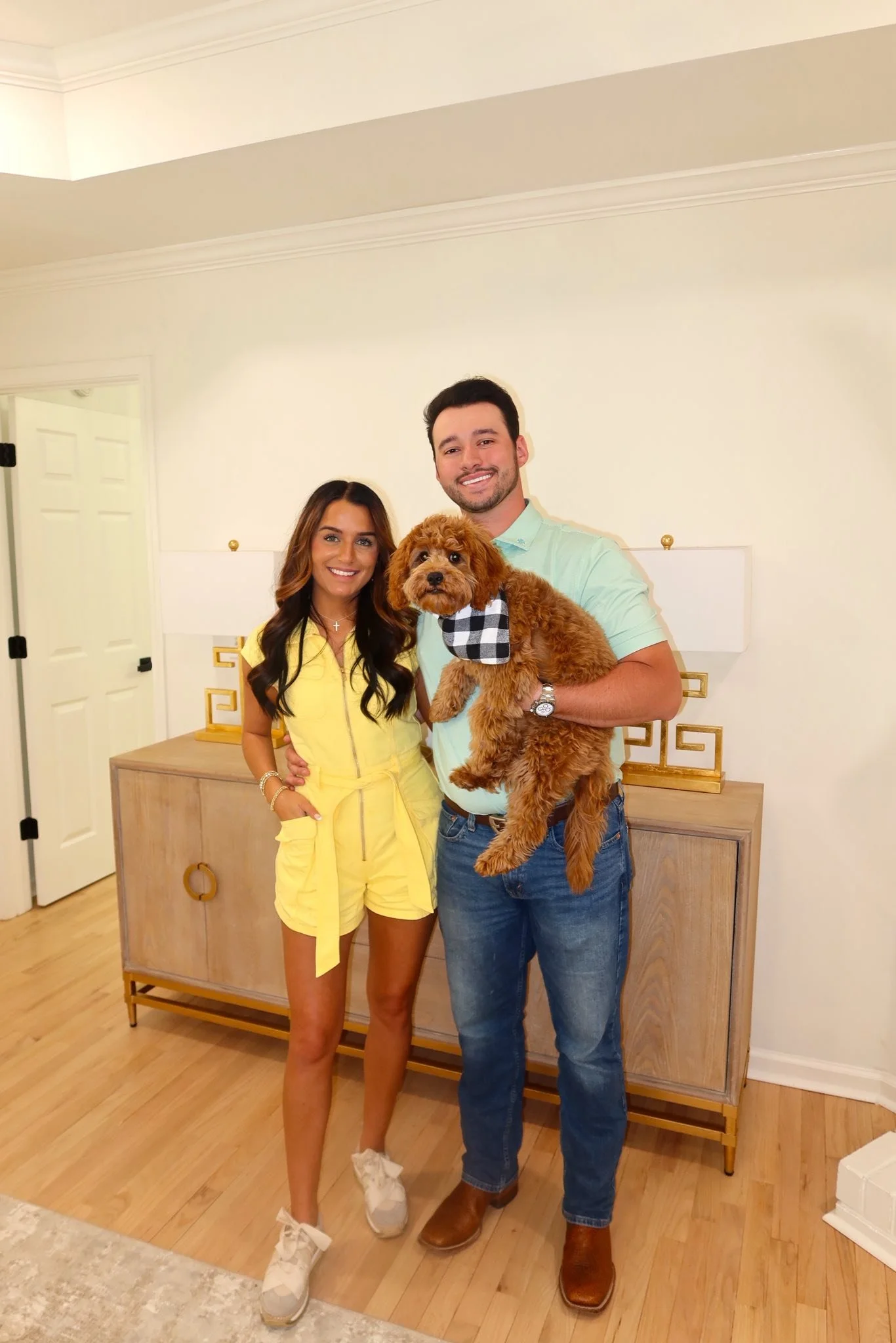 A smiling man and woman standing together, holding a small brown dog with a checked bandana, in a well-lit room with a wooden sideboard and decorative items.