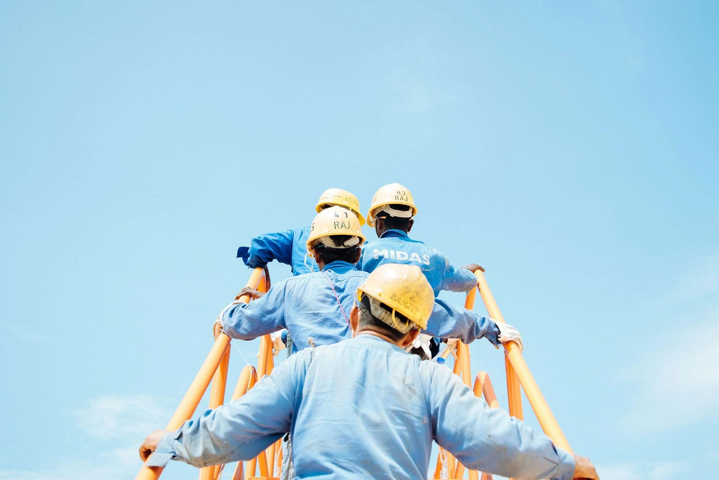 Construction workers wearing yellow helmets and blue uniforms climbing a ladder against a clear blue sky.