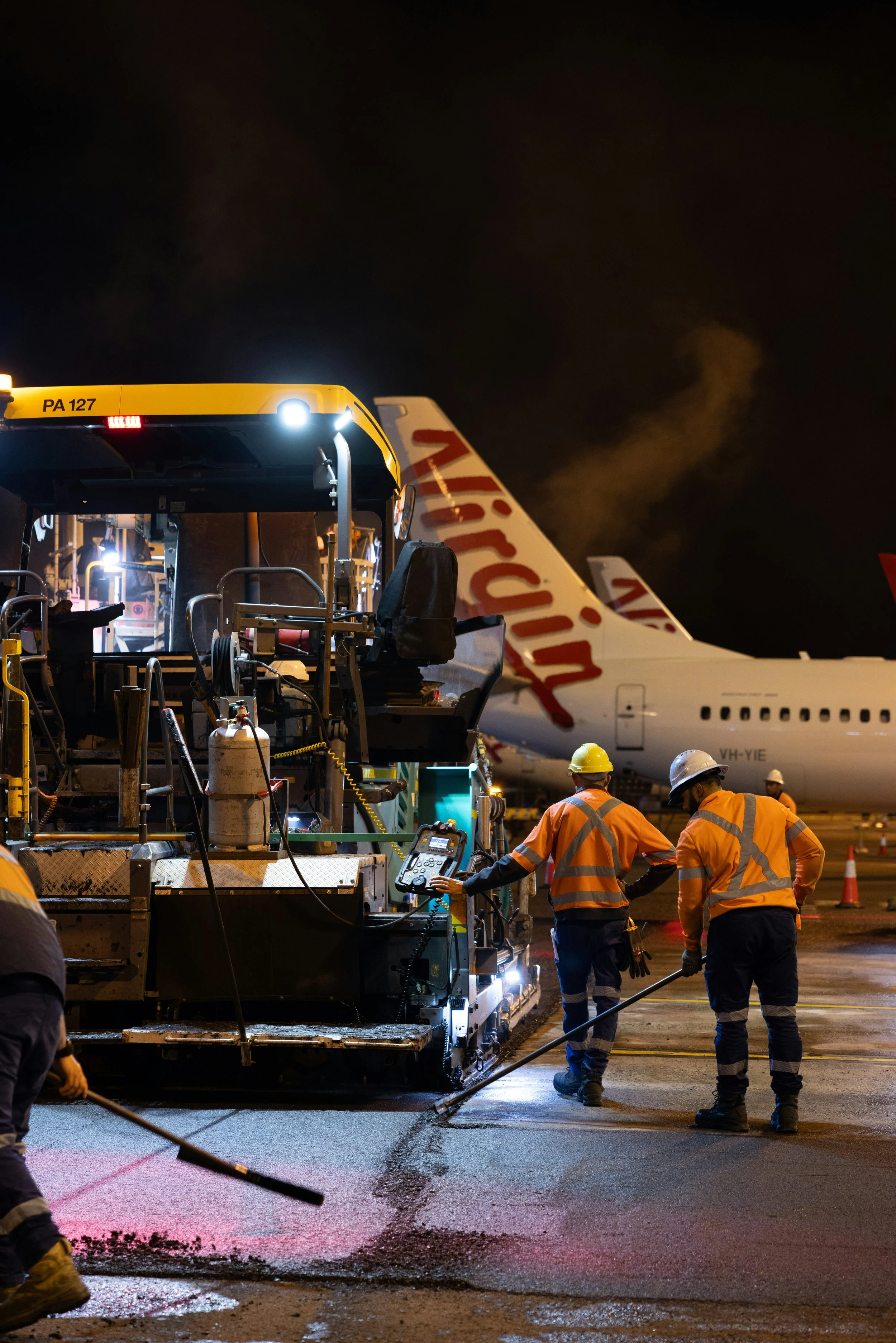 Nighttime airport runway workers paving freshly laid asphalt with machinery and workers in high visibility clothing and helmets