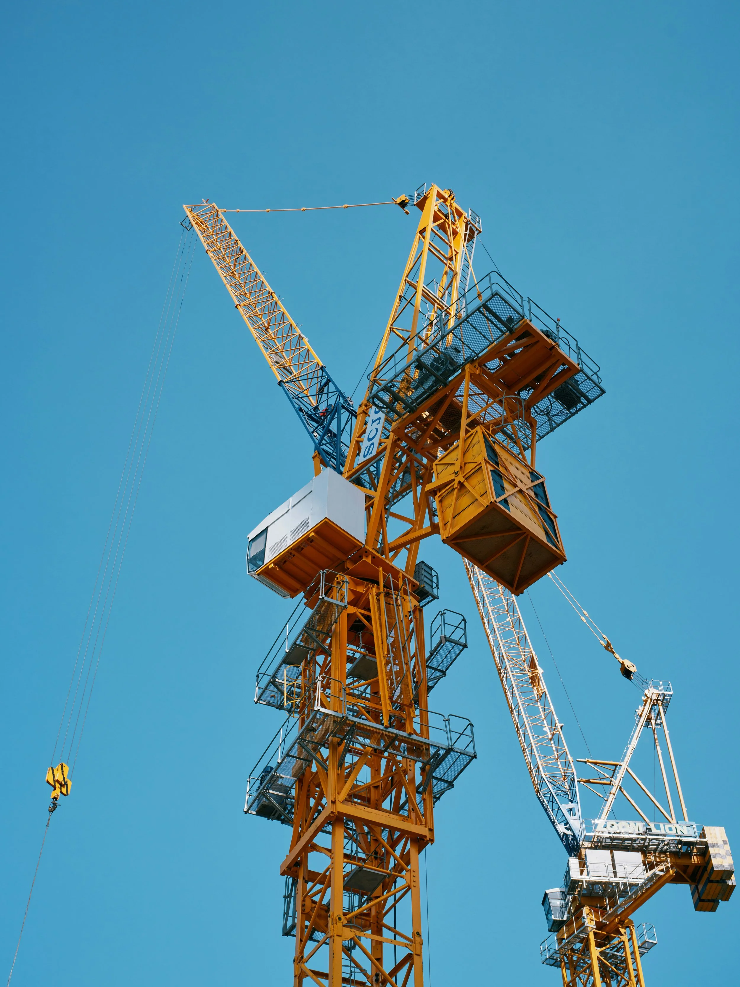 A yellow construction crane against a clear blue sky with some structures and parts of other cranes visible in the background.