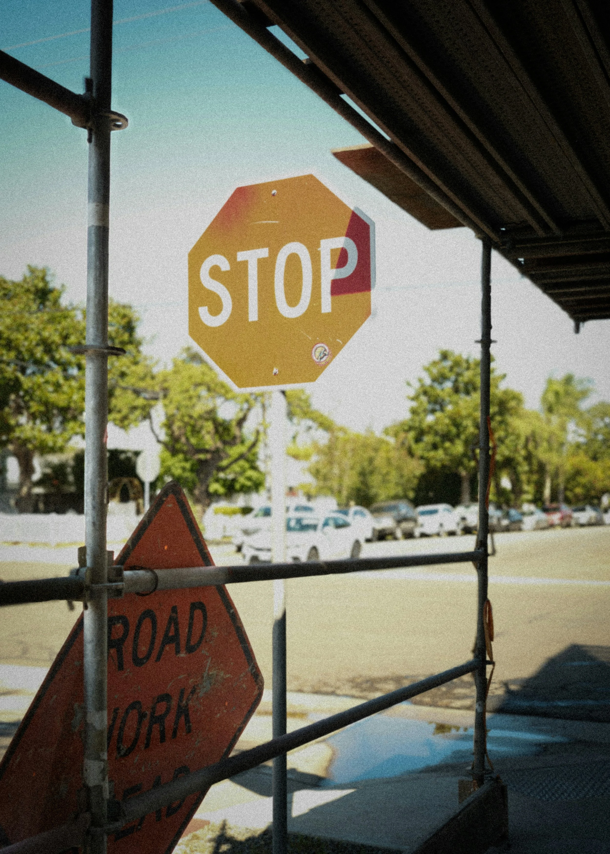 A yellow octagon-shaped stop sign with white letters is on a pole, partially covered by barriers. A red road work sign is on the ground below. In the background, trees and parked cars are visible, and the sky is clear.