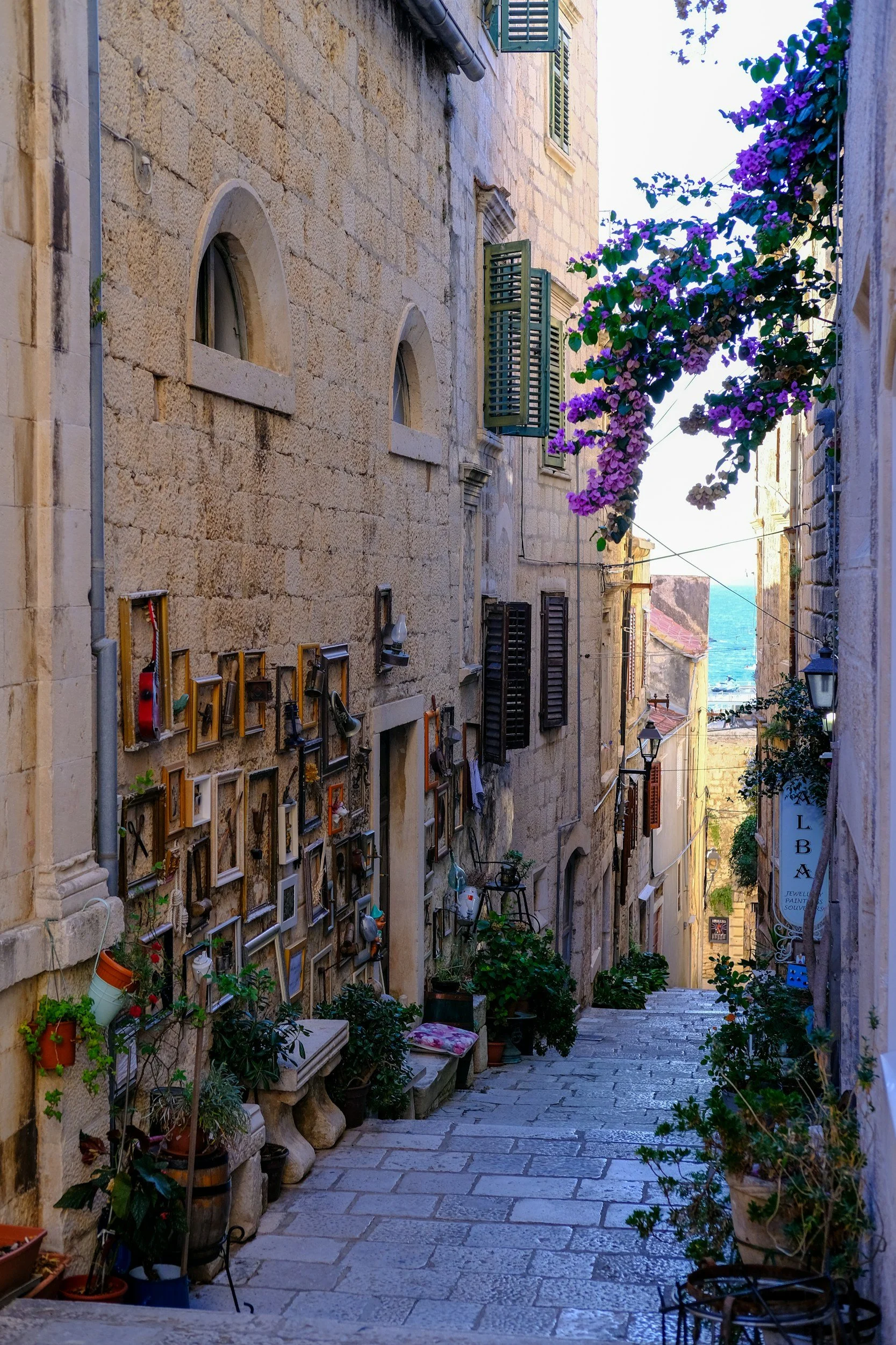 A narrow stone alleyway in a European city with flowering bougainvillaea and potted plants, showing buildings with shutters and small windows, and the sea visible in the distance.