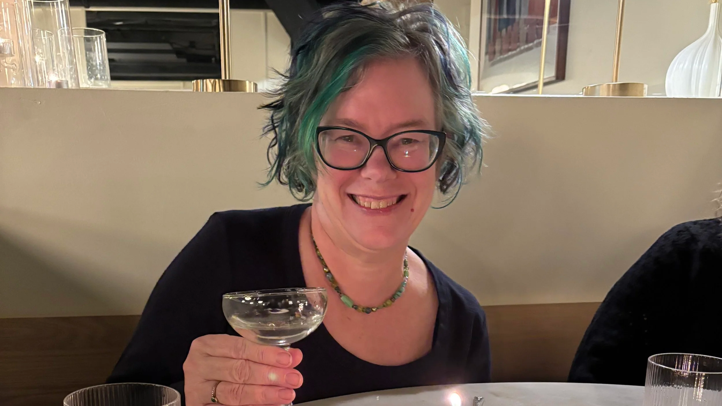 A woman with short, wavy green and blue hair, wearing glasses, a black top, and a beaded necklace, holding a coupe glass, smiling at a dinner table.