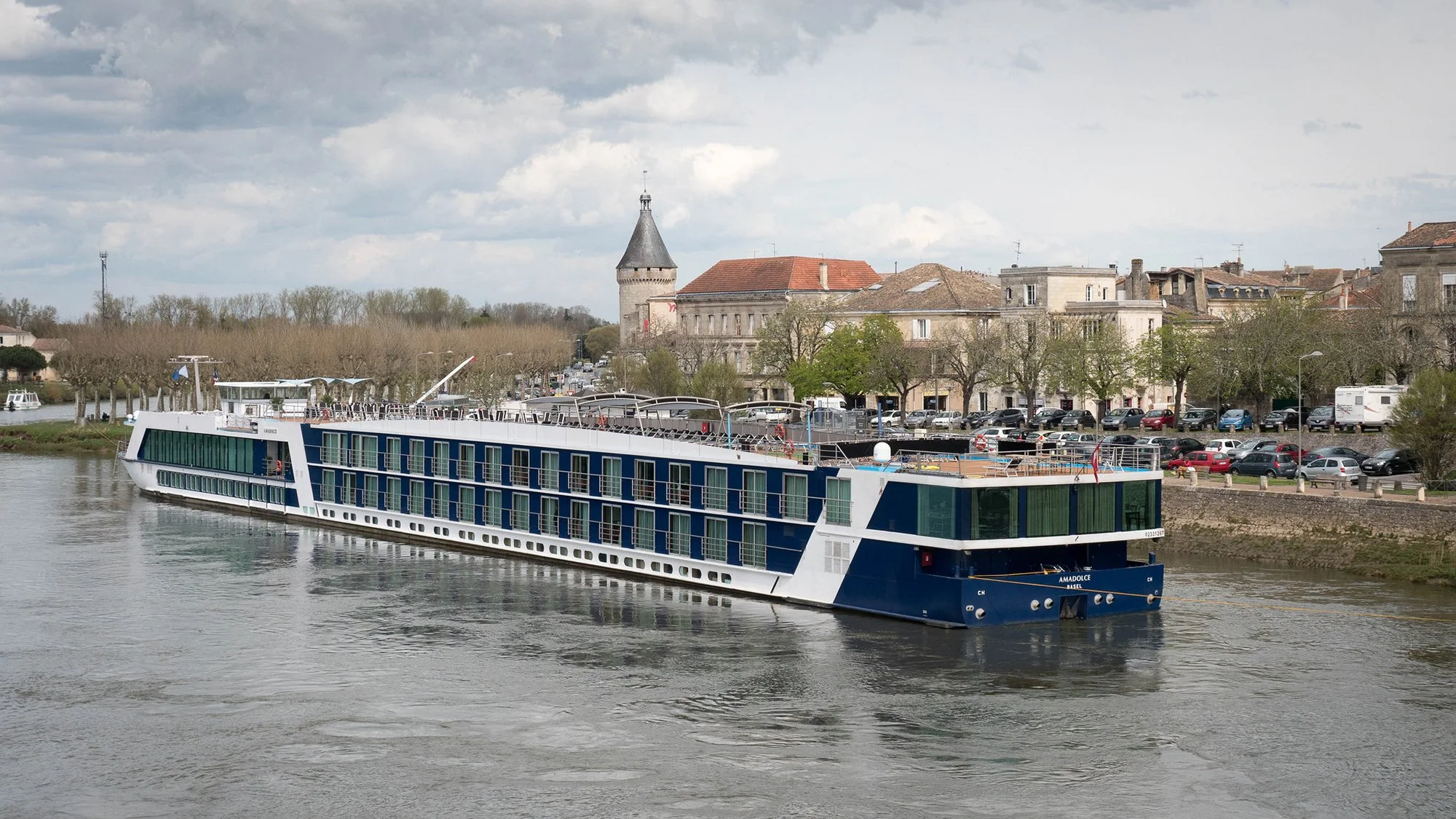 A modern, multi-level passenger cruise boat sailing on a river with a town and historic buildings in the background. Trees line the riverbank, and there are parked cars along the street. The sky is partly cloudy.