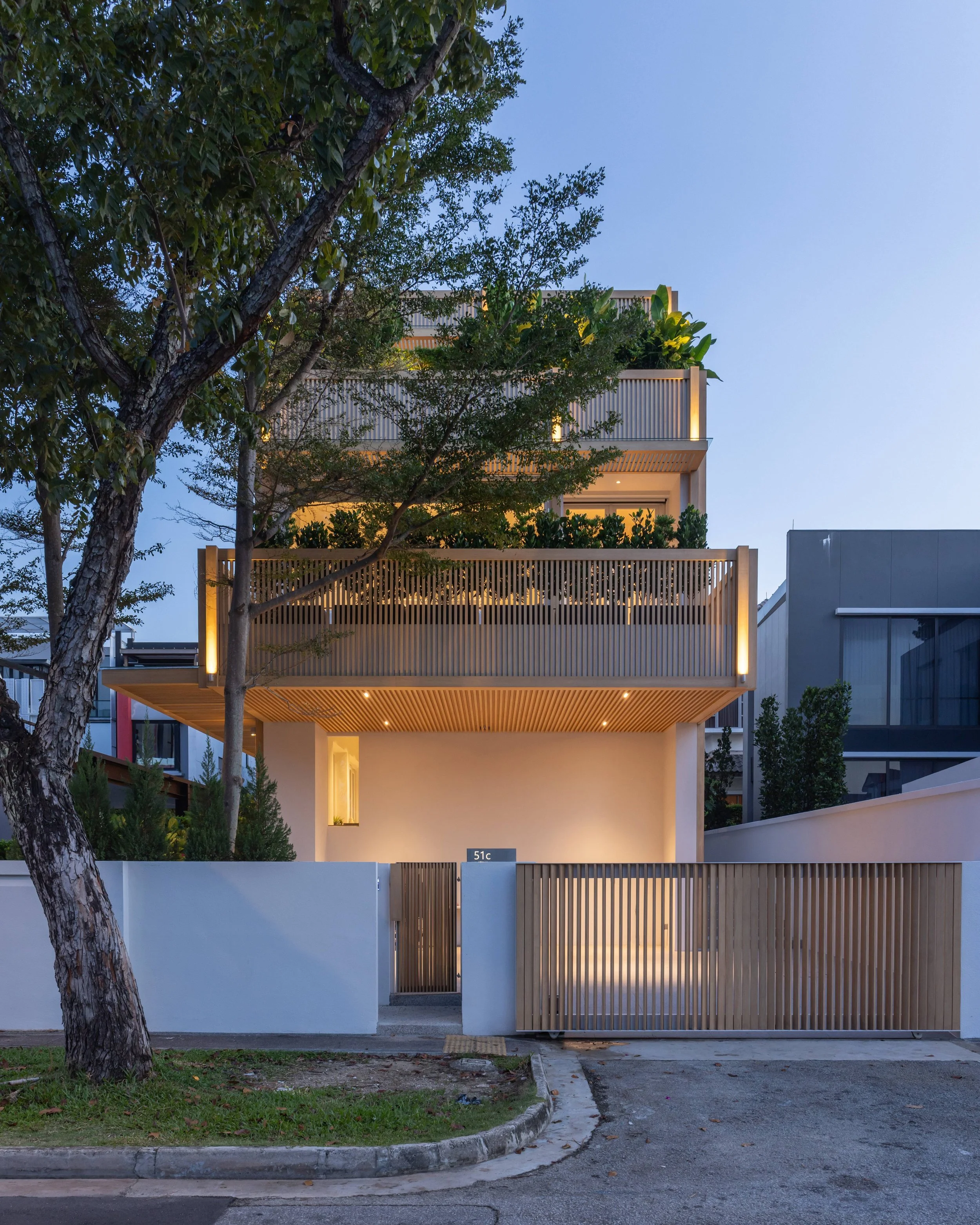 Modern multi-story house with a white and wooden exterior, surrounded by greenery and a white fence, with outdoor lighting at dusk.
