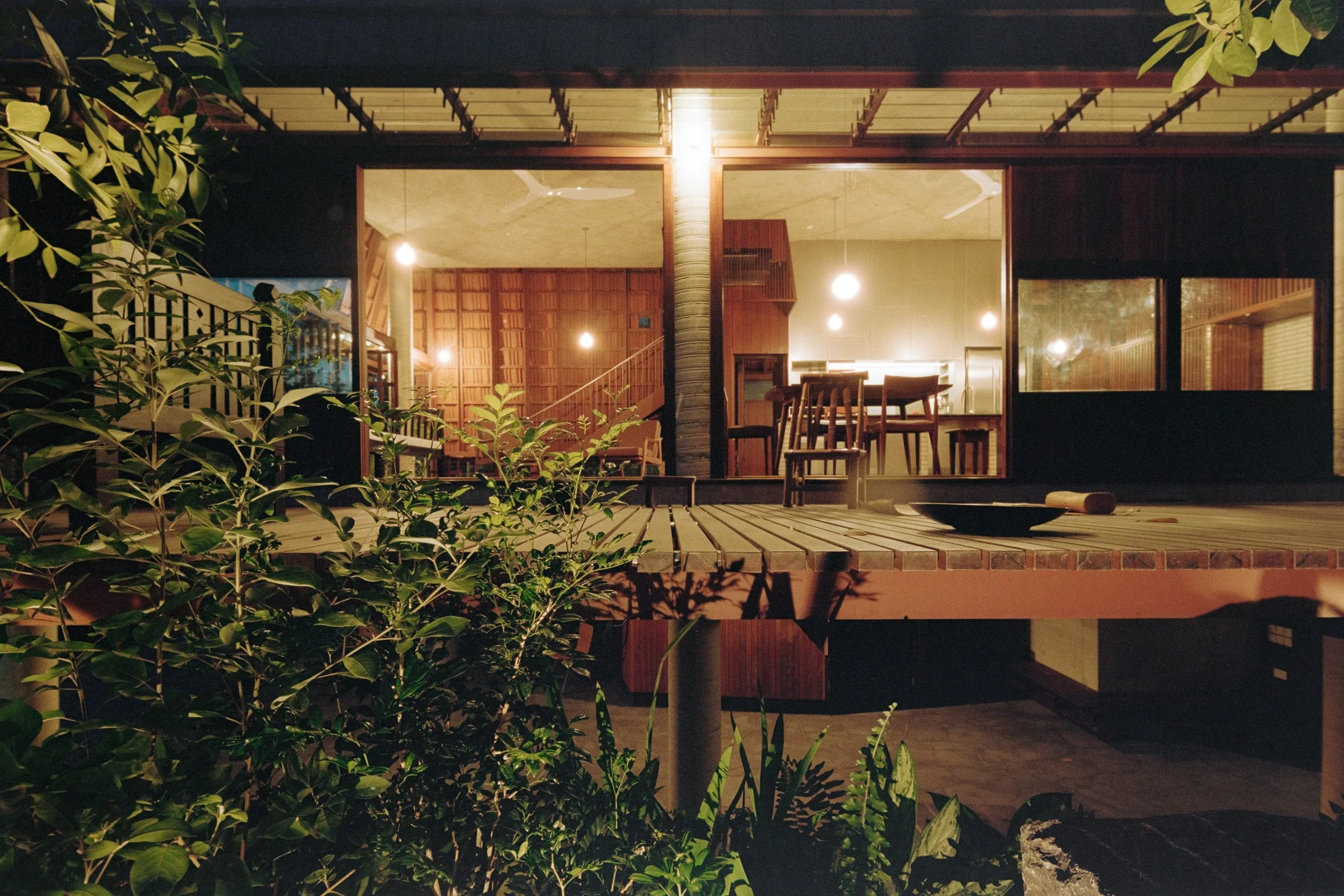 A cozy, well-lit interior viewed through large glass windows at night, with a wooden dining table and chairs, and a staircase inside. Outside, a wooden deck with some chairs, a bowl, and greenery in the foreground, with warm lighting creating a serene atmosphere.