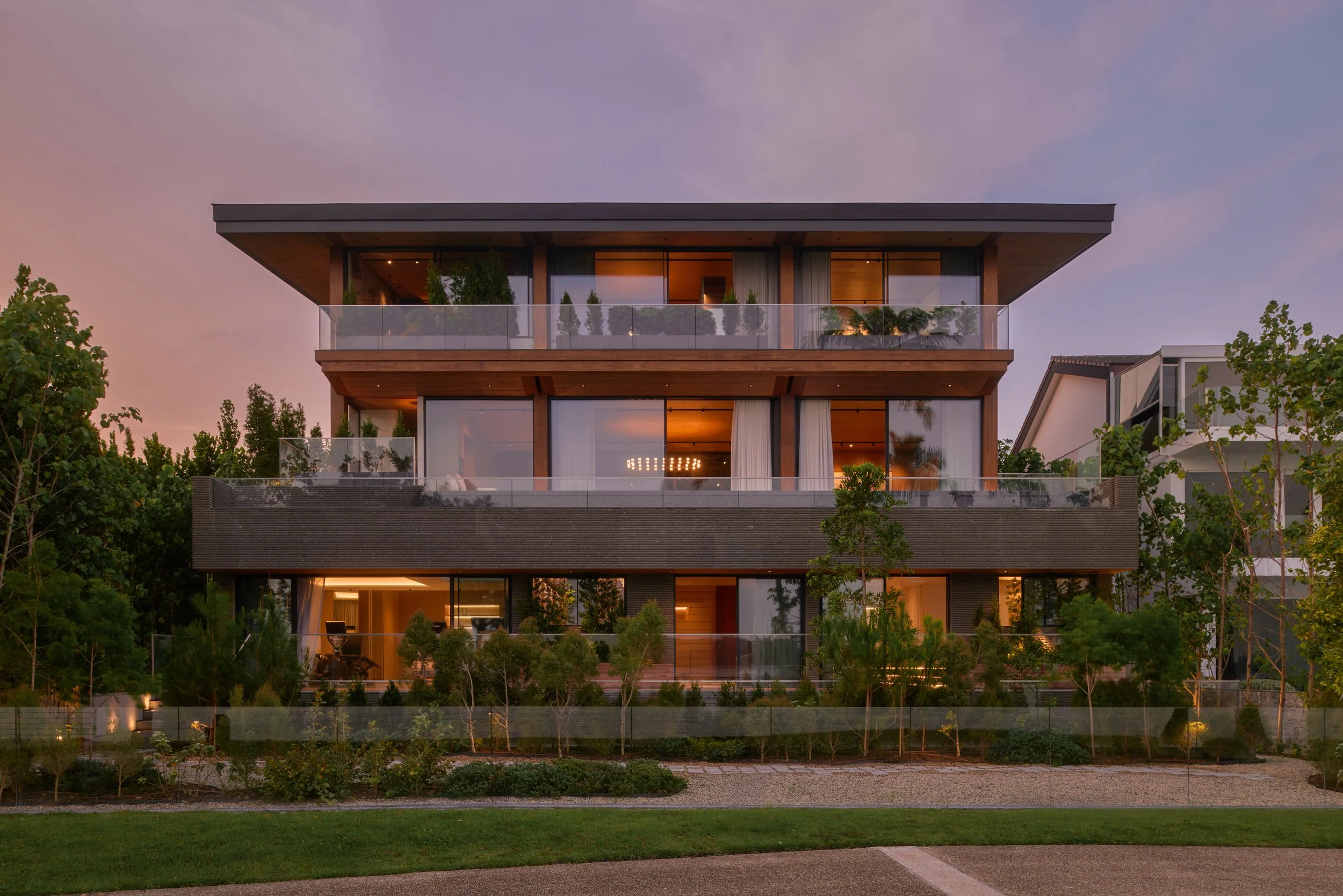 Modern three-story residential building with large glass windows and balconies, surrounded by greenery, during sunset.