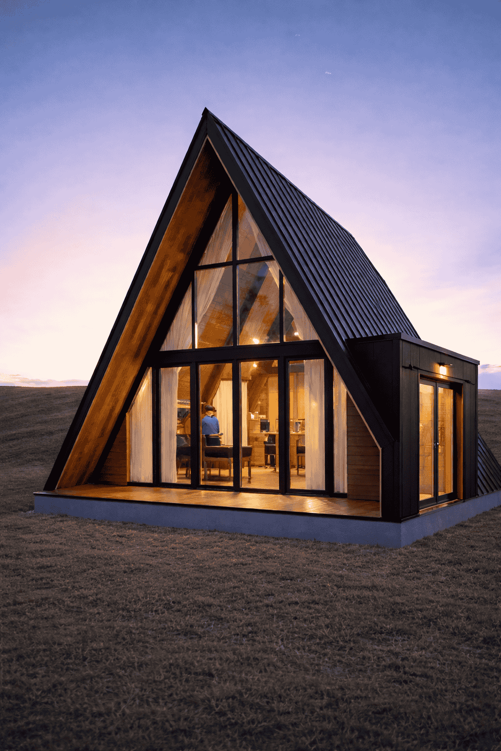 Modern A-frame house with large glass windows, black exterior, located in a grassy field at sunset.