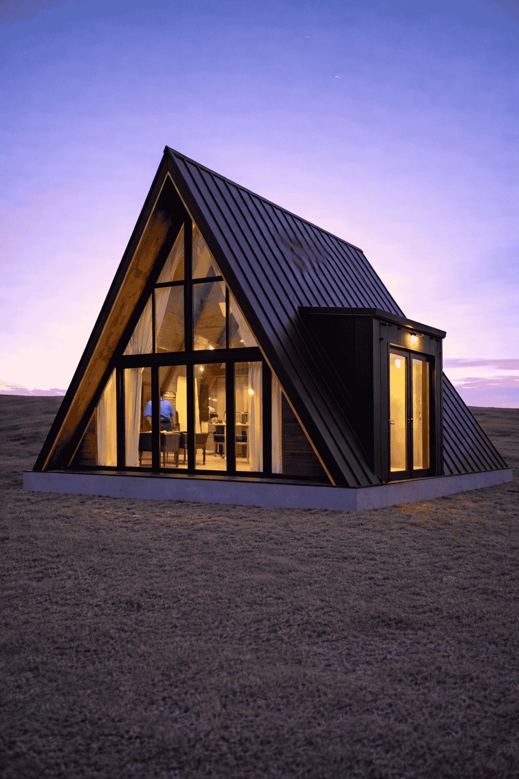 A modern A-frame house with large glass windows and a metal roof, glowing warmly inside at dusk, situated on a grassy field.