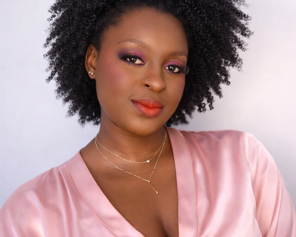 Close-up portrait of a woman with natural curly black hair, wearing pink lipstick, purple and pink eye makeup, gold earrings, layered necklaces, and a silky pink blouse against a light background.