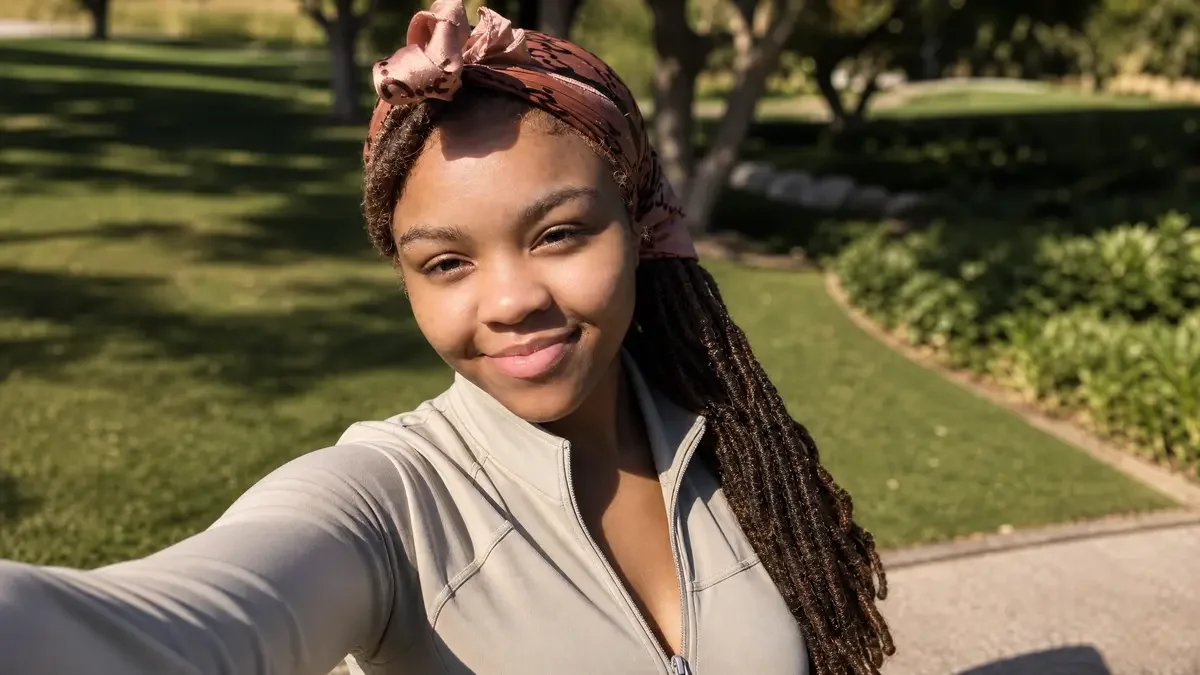 A young woman with long braided hair, wearing a brown headscarf and a light-colored jacket, smiling outdoors on a sunny day with trees and a grassy park in the background.