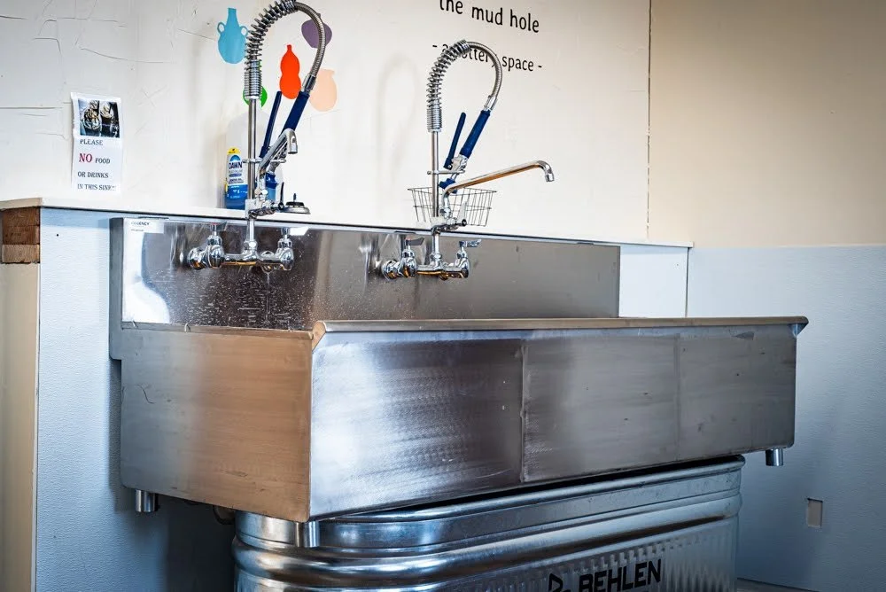 Industrial stainless steel laundry sink with two faucets and sprayers, set against a wall with signs and decorations.
