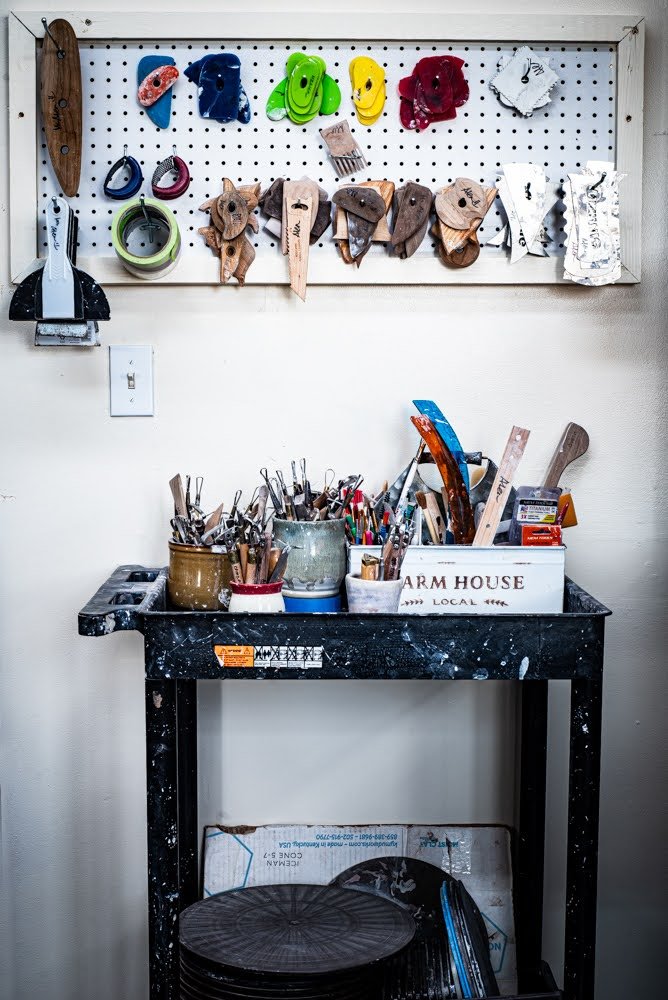 Workshop space with a black work table filled with paintbrushes and tools in buckets. Mounted pegboard with colorful hooks, saws, and other tools above the table.
