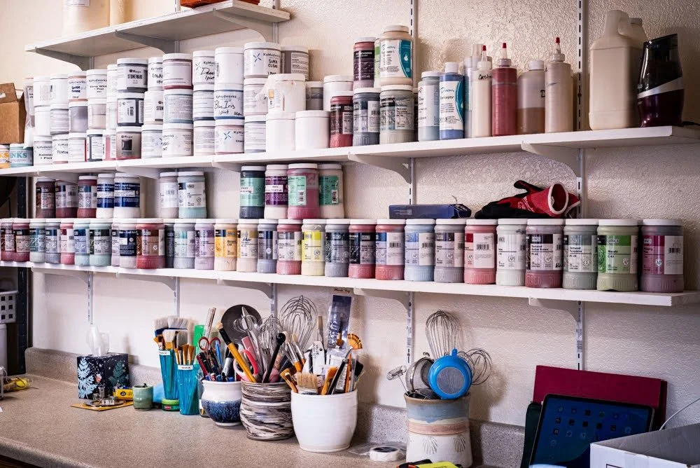 Shelves filled with various bottles of paint, paintbrushes, and art supplies in jars and containers in an art studio workspace.