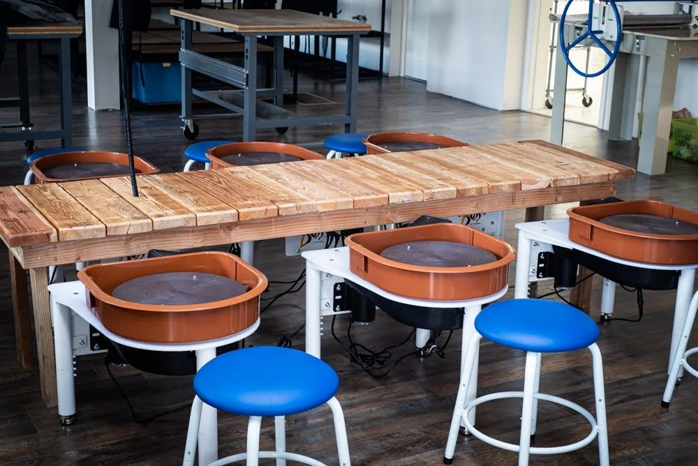 A ceramics studio workspace with pottery wheels, a wooden work table, and stools, set on a hardwood floor.