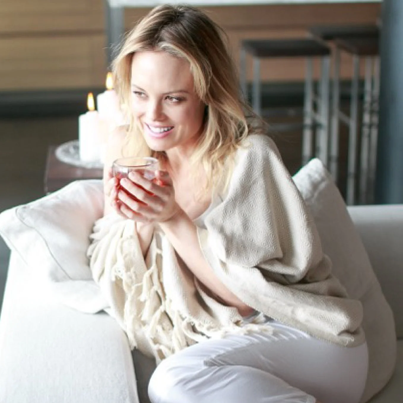 A woman with blonde hair sitting on a sofa, holding a glass of red wine, smiling, with candles in the background.