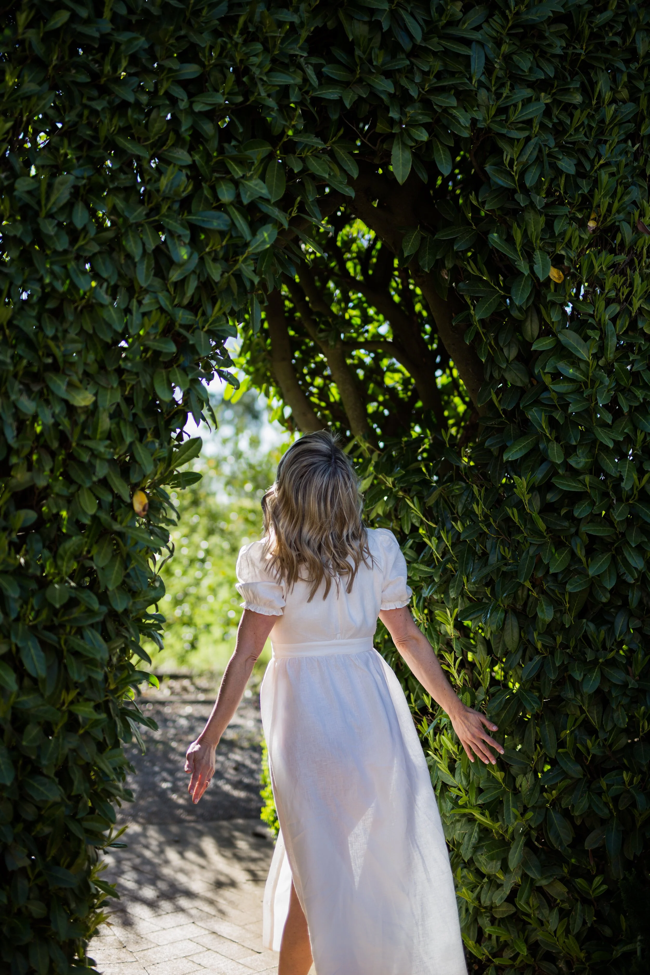 A woman with blonde, wavy hair wearing a white dress walks through a lush green hedge in an outdoor setting, sunlight filtering through the leaves.