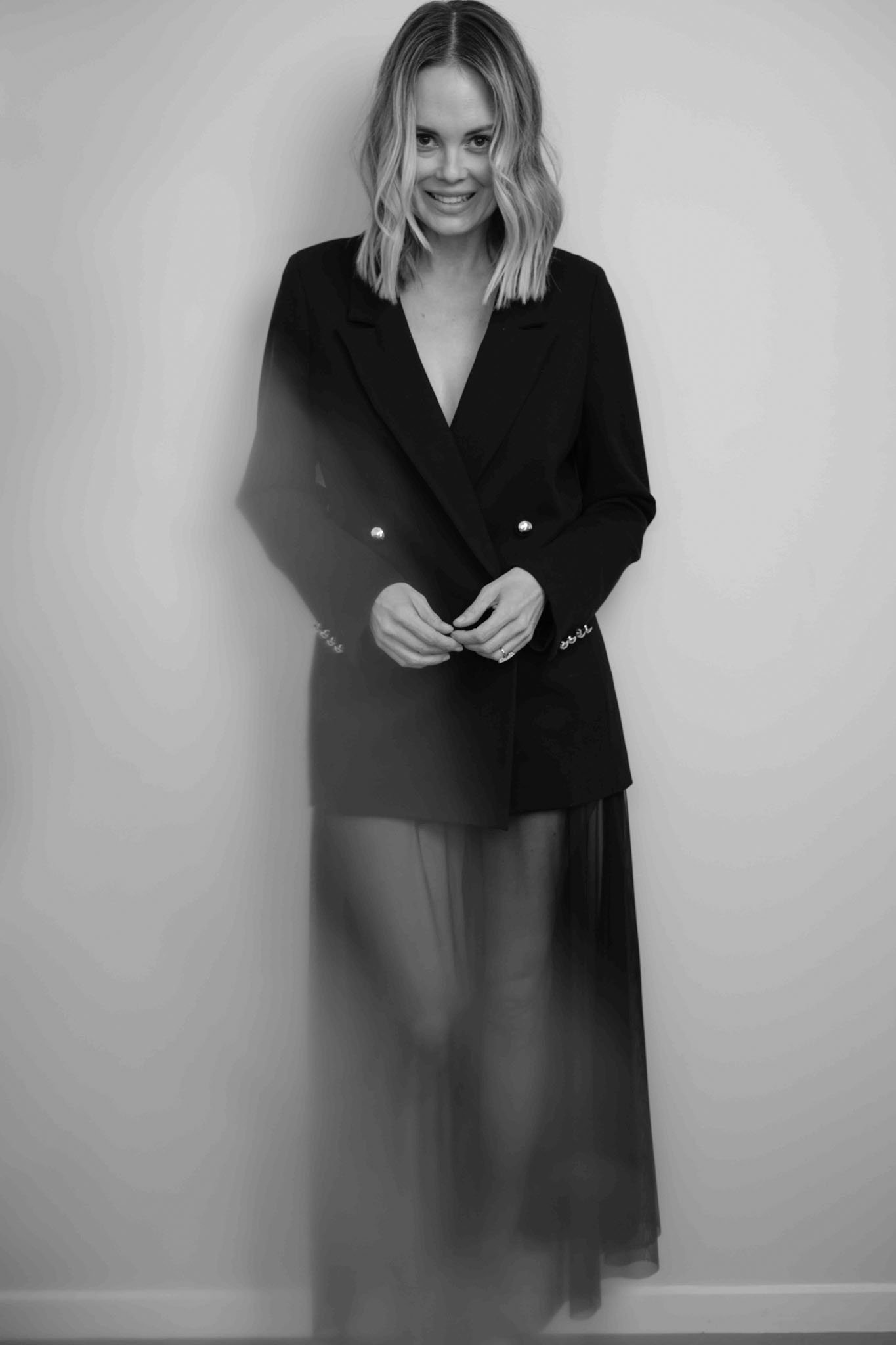 Black and white photo of a woman with shoulder-length wavy hair smiling, wearing a dark blazer over a dress or skirt with a sheer overlay, standing against a plain wall.