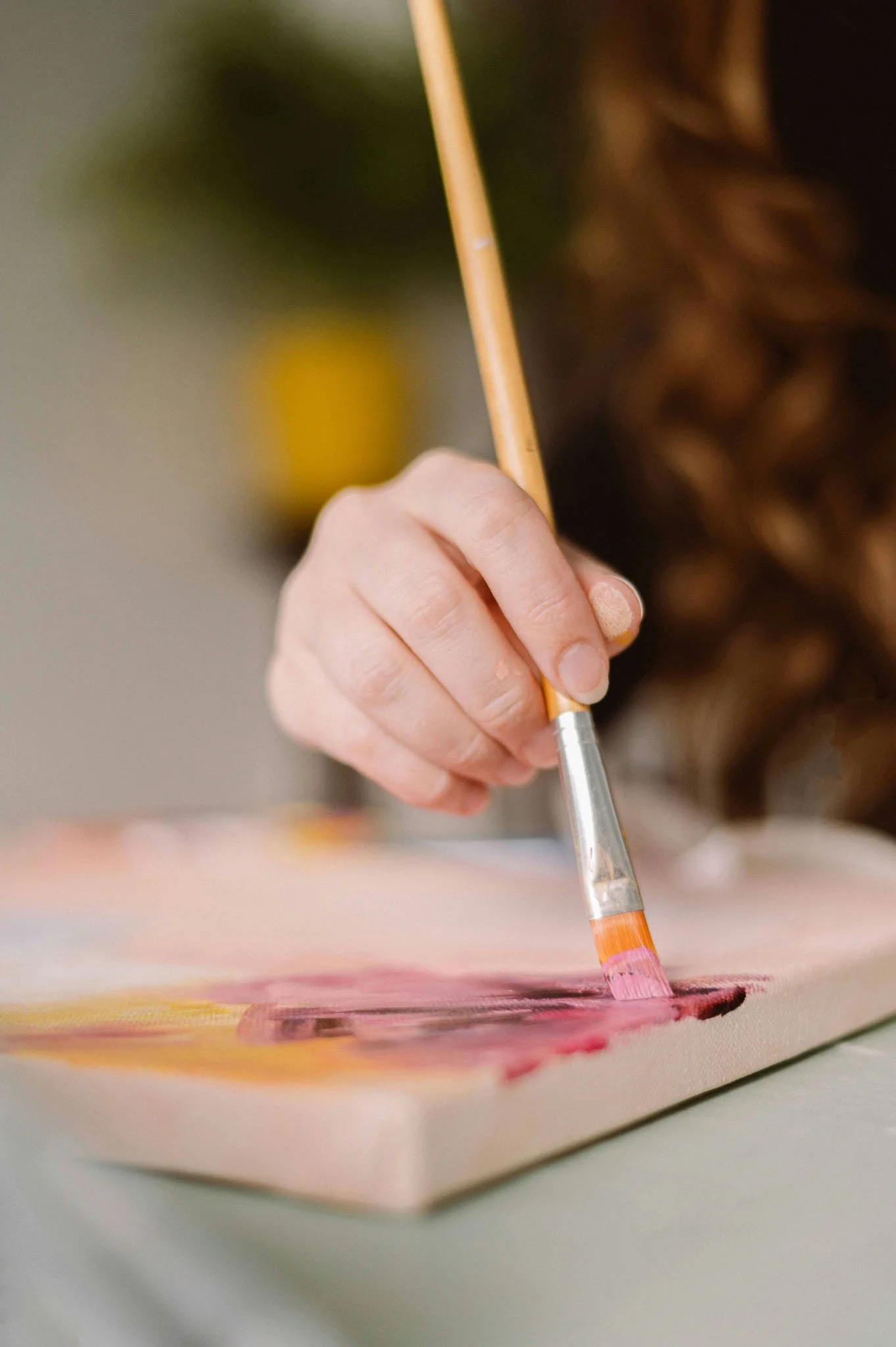 Close-up of a person holding a watercolor brush, painting colorful strokes on paper. The person's face is partially visible with brown curly hair, focused on their artwork.