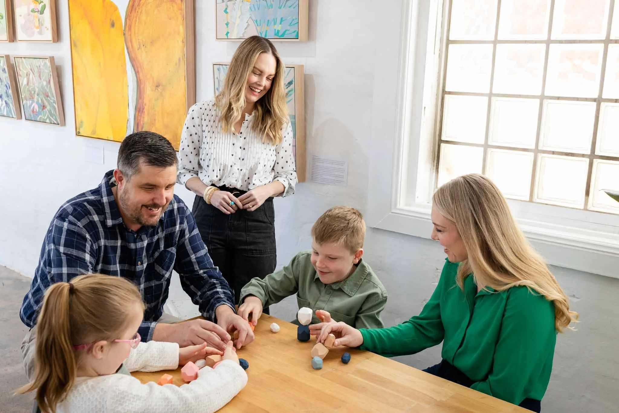 A group of children and adults playing with rocks at a table in an art gallery. The walls are decorated with colorful abstract paintings and large windows.