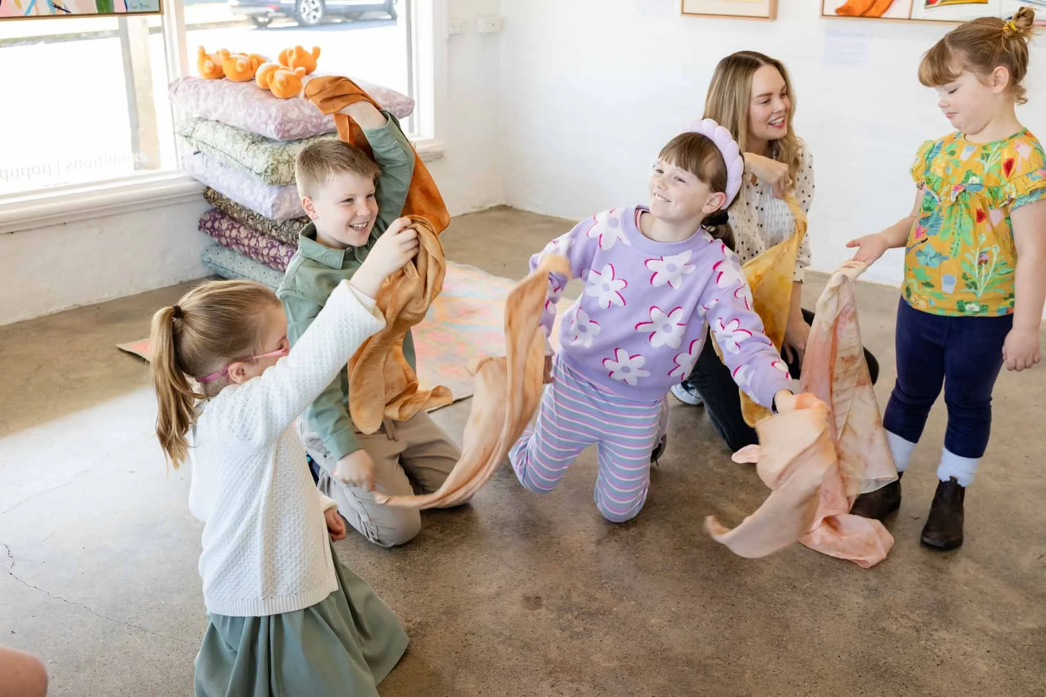 Children and a woman playing a game with a long cloth in a bright indoor space.