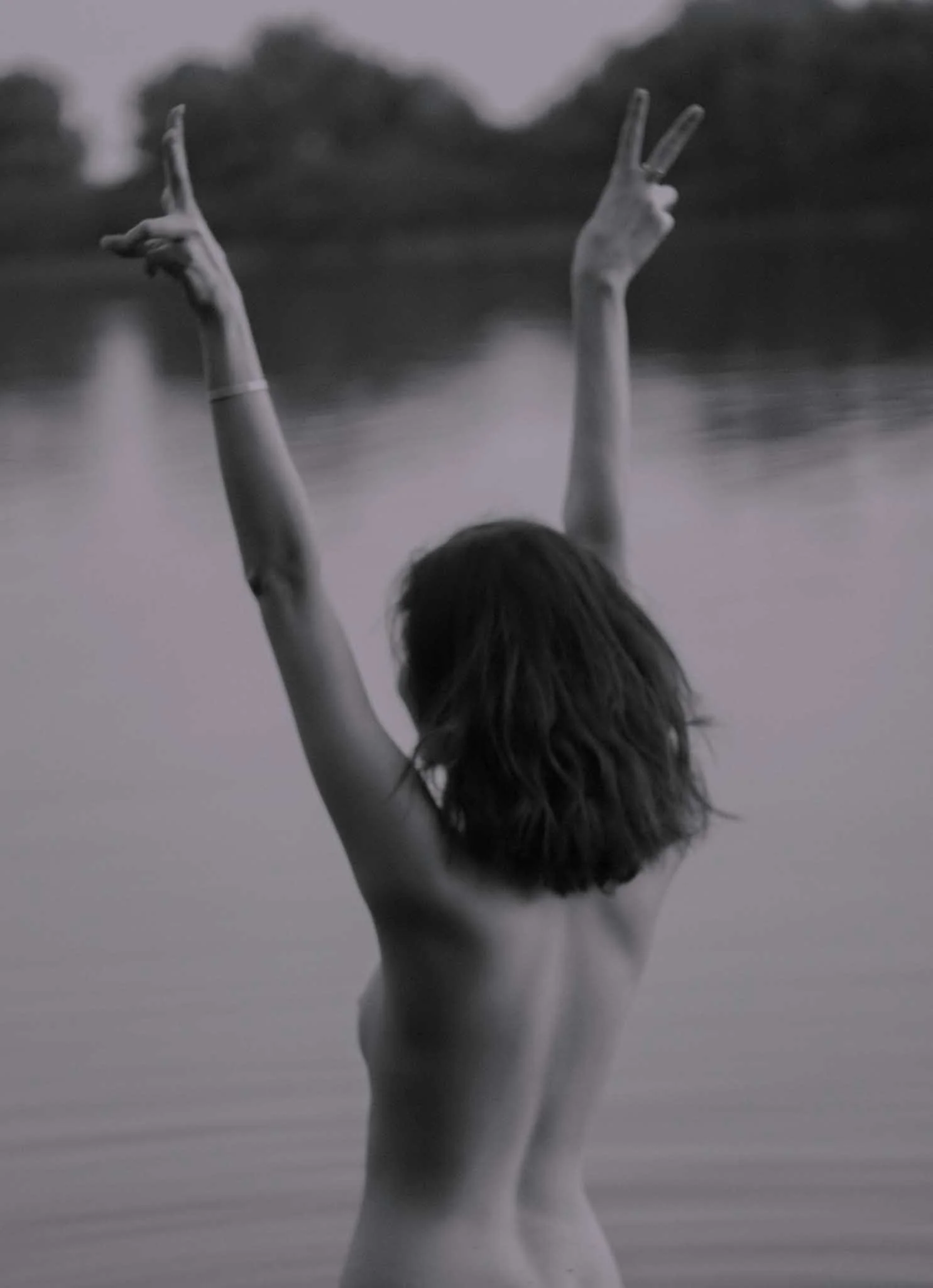 A black and white photo of a person with long hair seen from behind, raising their arms in the air, with arms bent and fingers forming peace signs, standing in shallow water with a blurred background of trees.