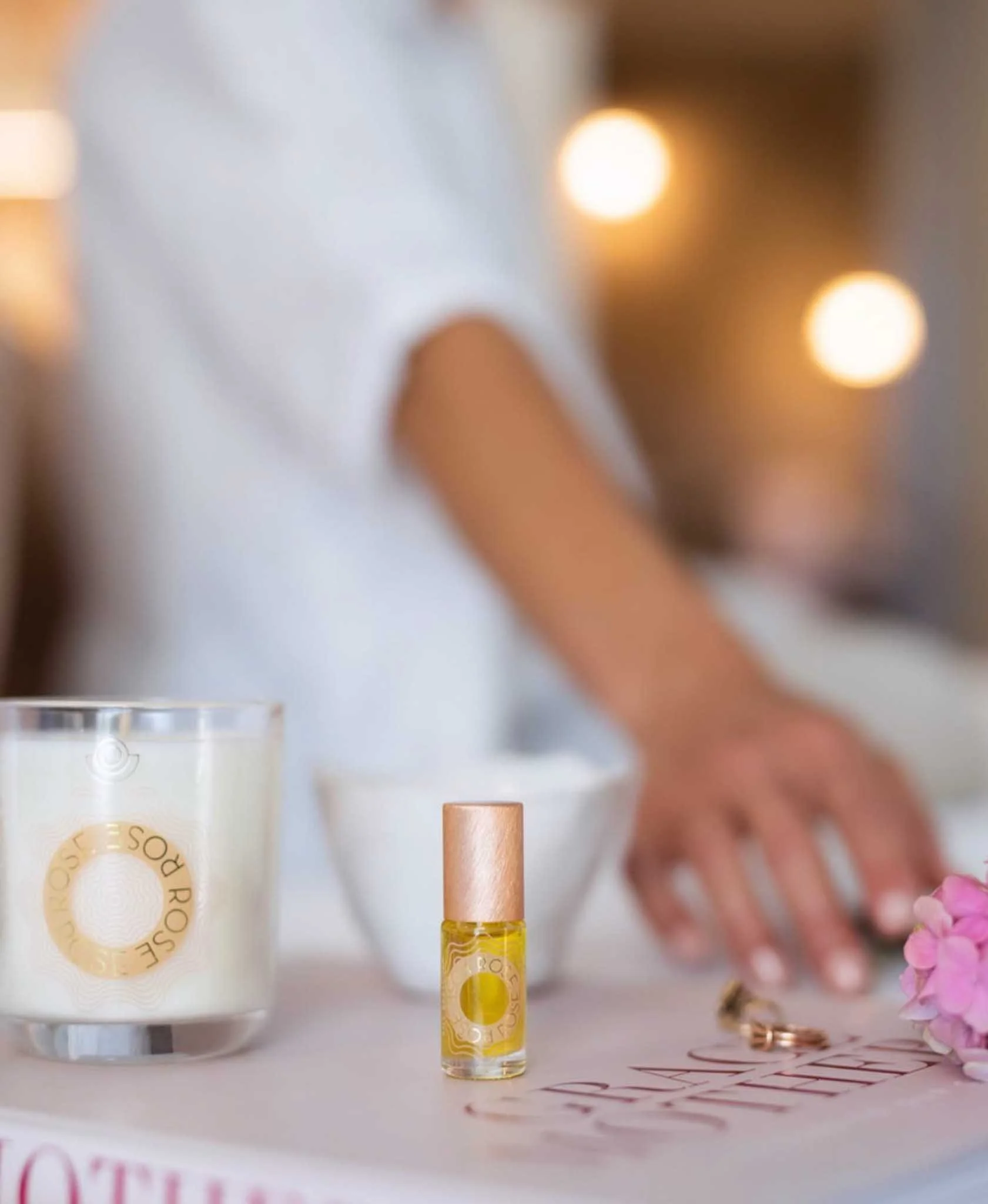 A small glass vial with yellow liquid and a gold lid, surrounded by a larger box, pink flowers, and a piece of paper with handwritten notes, on a white surface.