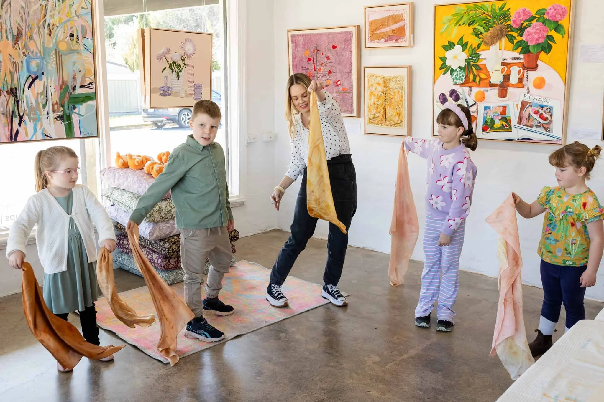 Children and an adult standing on a colorful rug in an art gallery, holding fabric pieces, with framed abstract and floral artworks on the walls.