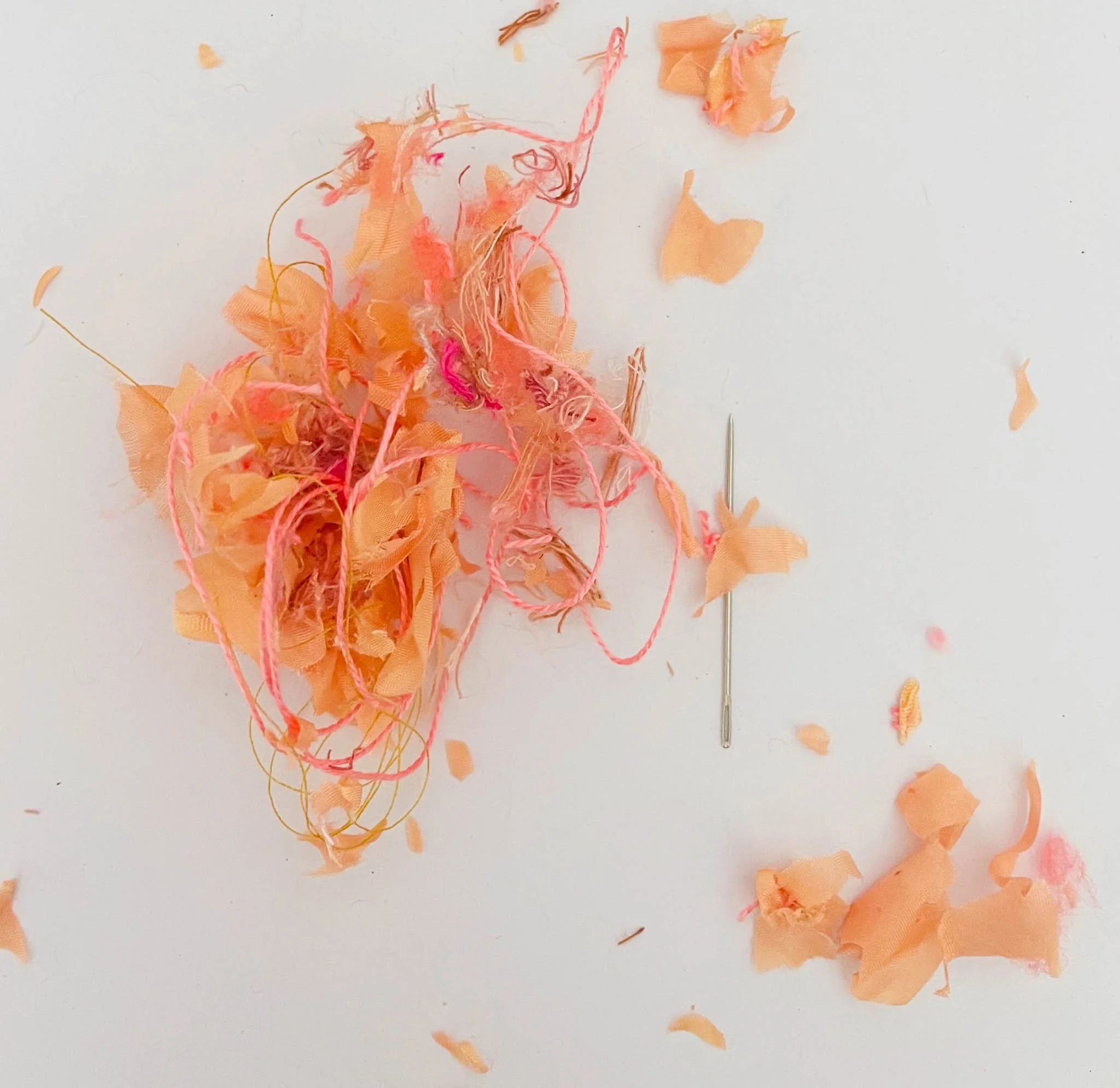 A sewing needle lying on a white surface surrounded by orange and pink fabric scraps and threads.