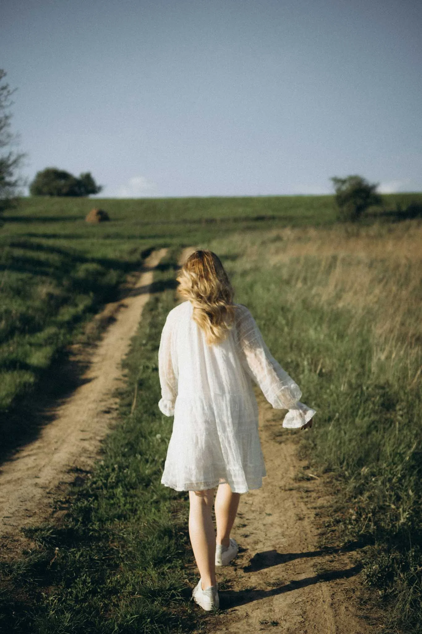 A woman walking away along a dirt path through a grassy field with trees in the distance under a blue sky.