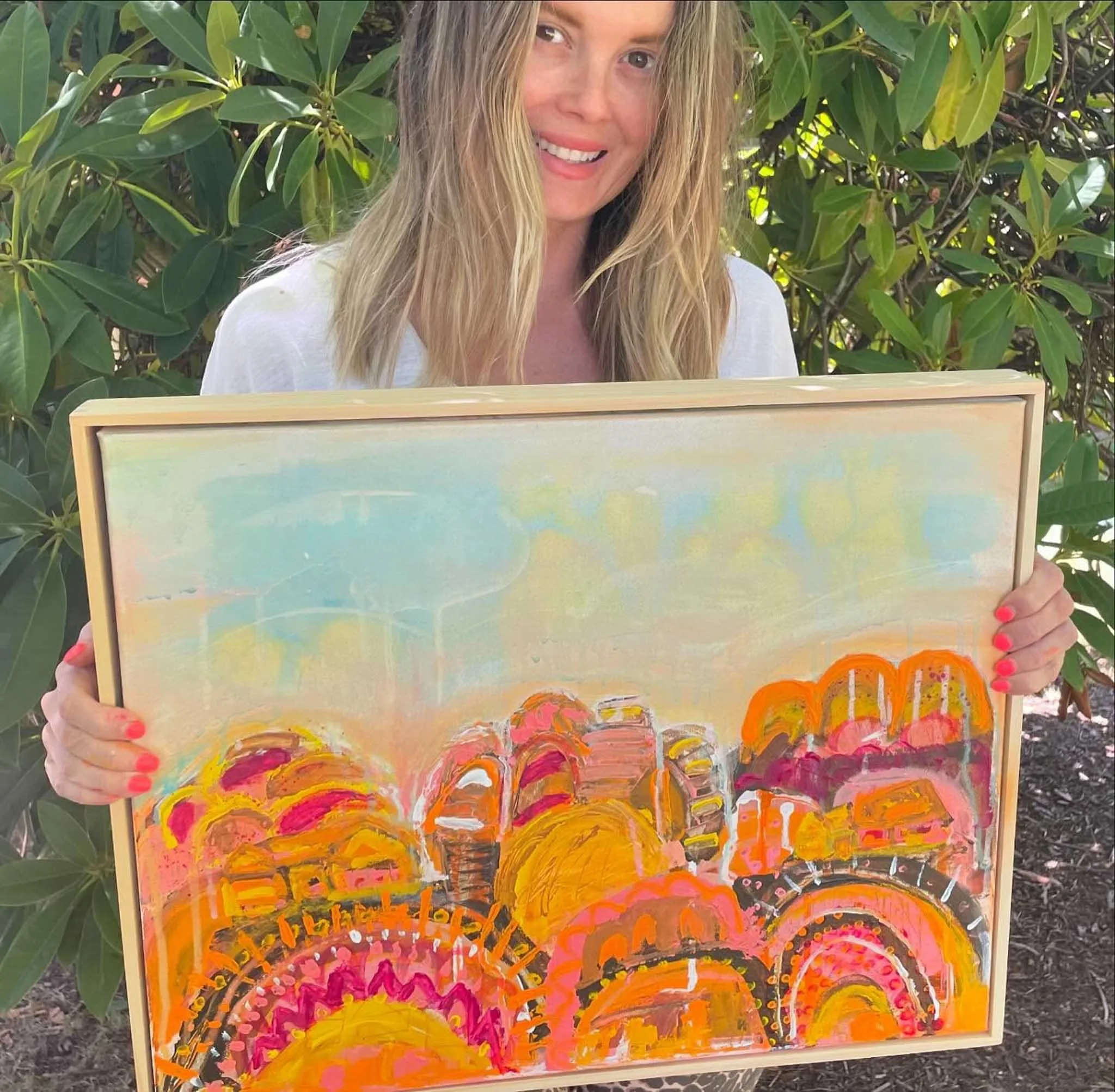 Woman with long blonde hair and a white shirt holding a colorful painting with orange, pink, yellow, and black brushstrokes while standing outdoors in front of green leafy bushes.