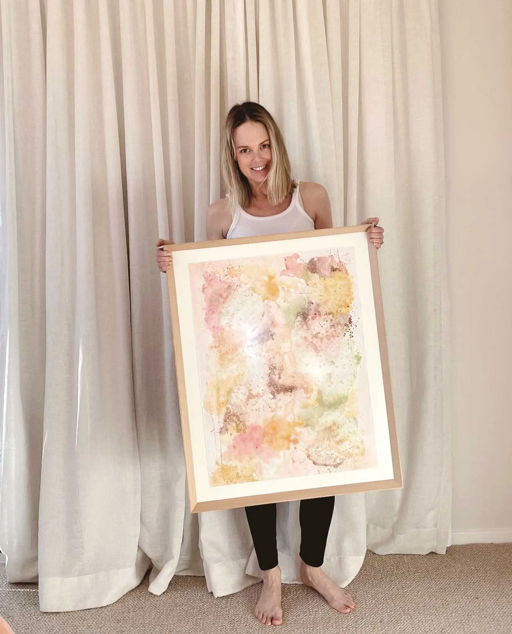 Woman with blonde hair, wearing a white tank top and black pants, holding a framed abstract watercolor artwork, standing in front of a beige curtain.