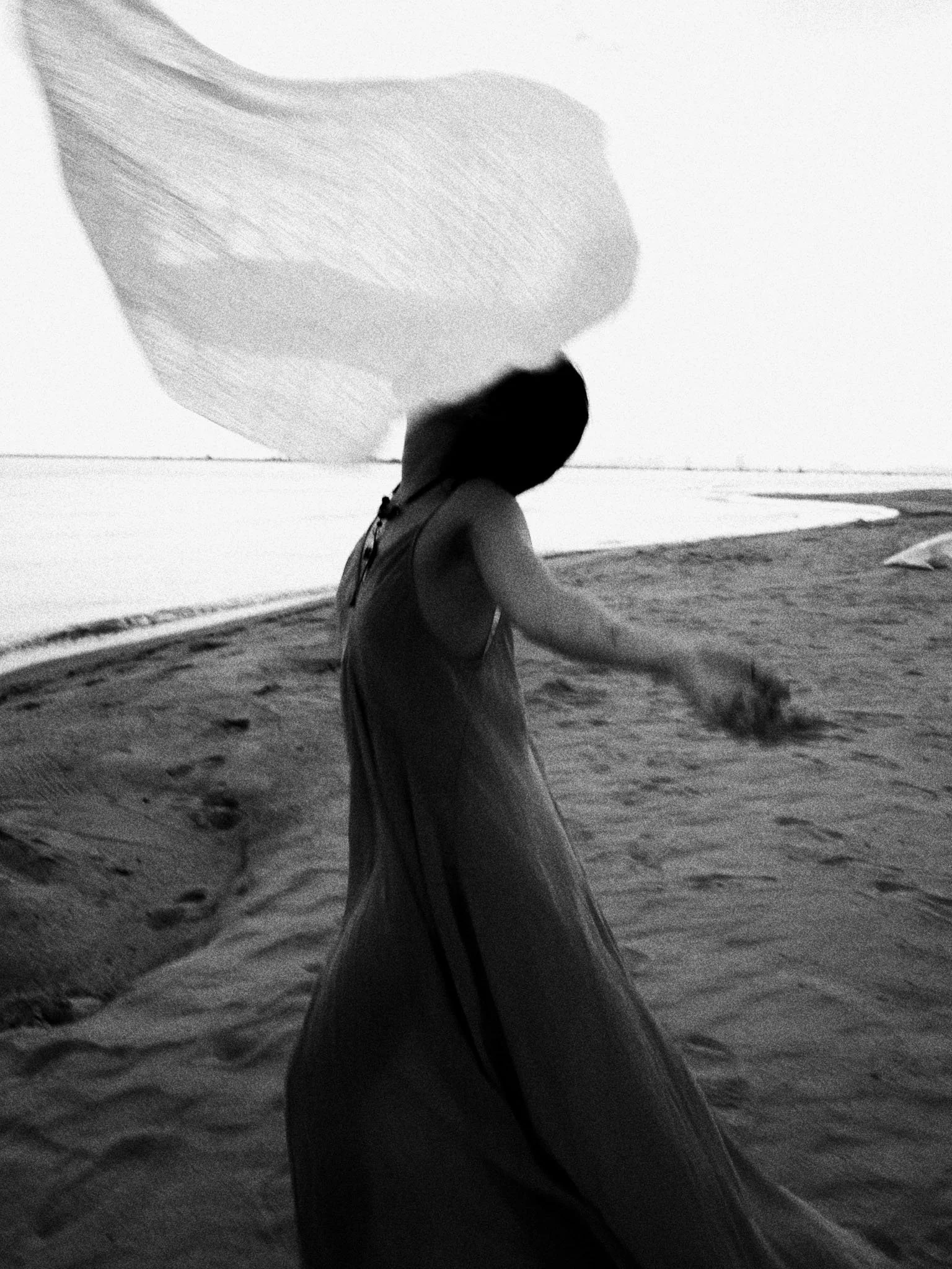 A person walking on the beach during sunset, seen from behind, with long hair, wearing a dress, and a large cloud or umbrella in the sky.