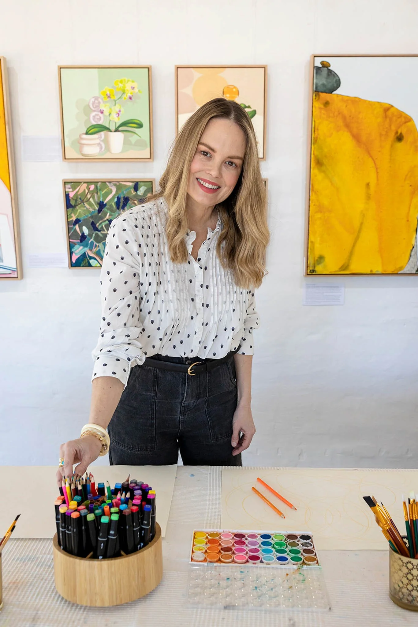 A woman with long blonde hair smiling at an art gallery or studio, standing behind a table with art supplies including markers, watercolors, and drawing tools, with colorful abstract paintings on the wall behind her.